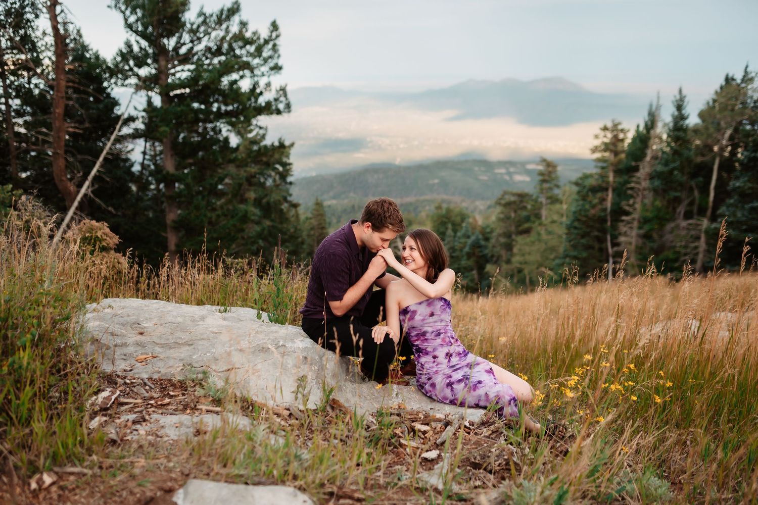 A couple sits on rocky terrain with mountains and trees in the background.