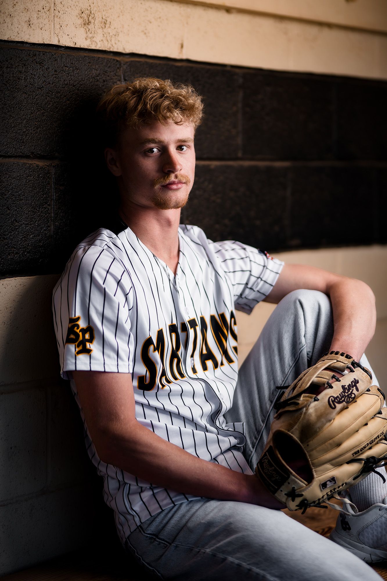 Baseball player in pinstripe uniform sits in dugout with glove during sports photography session.