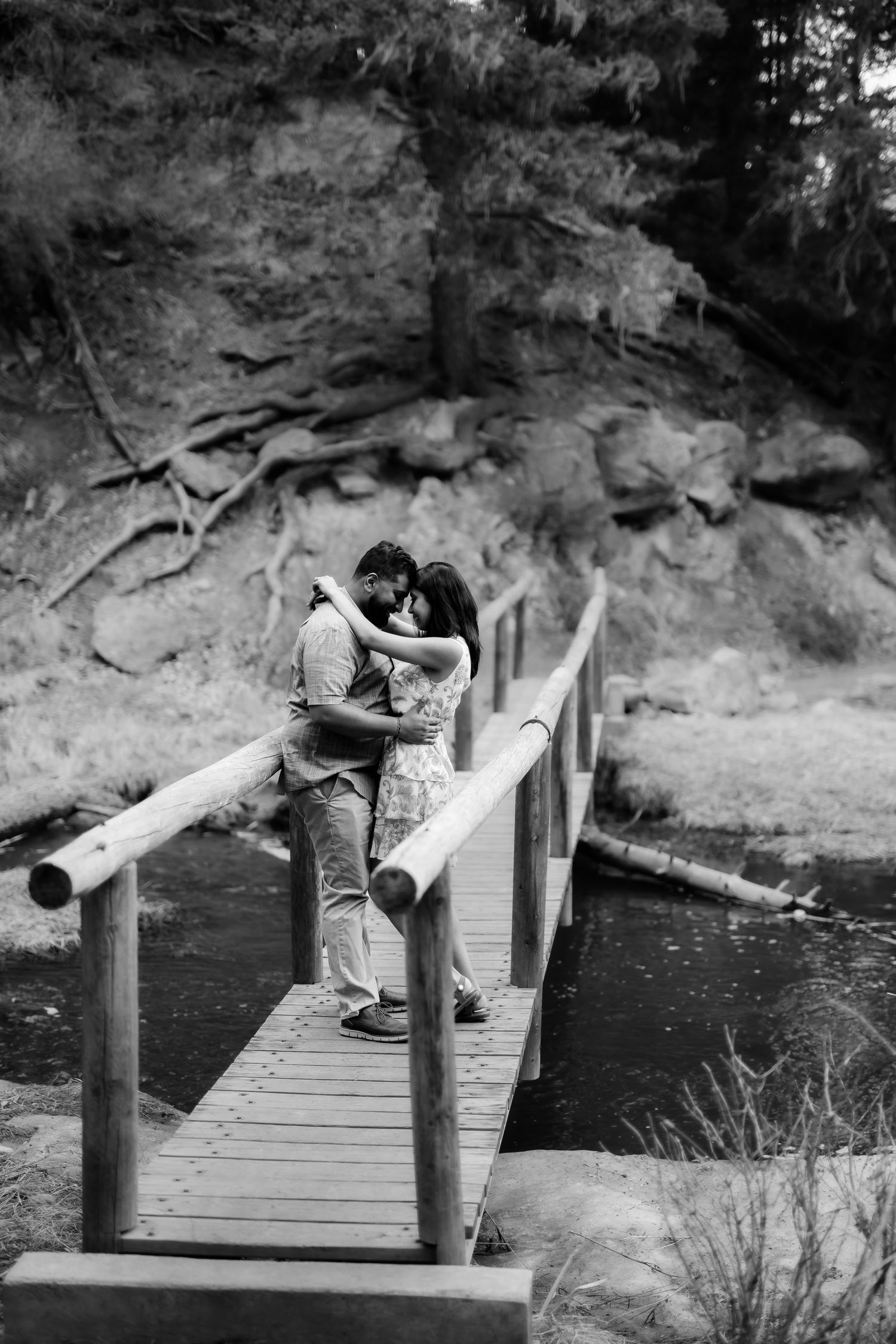 A romantic couple embracing on a wooden bridge over water with rocky cliffs in the background.