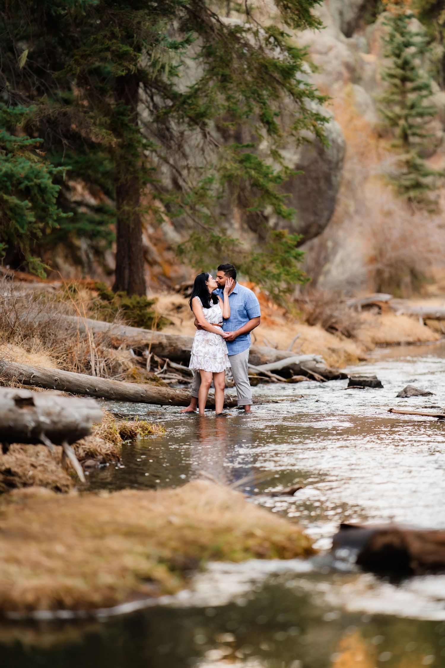 A romantic couple embraces while standing in a shallow stream surrounded by rocky cliffs and evergreen trees.