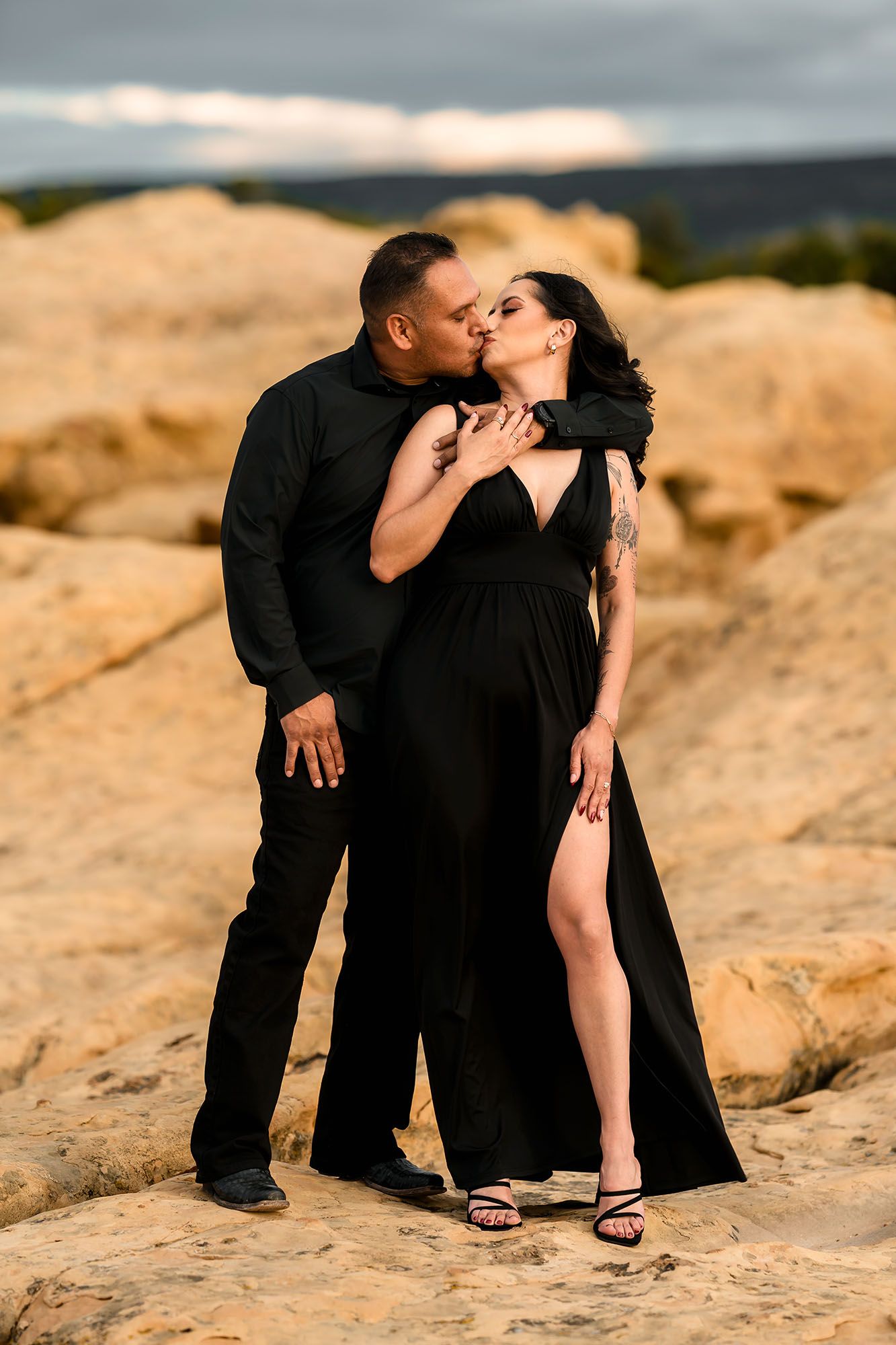 Couple in matching black outfits kissing on golden rocks at sunset, woman in elegant slit gown with tattoos.