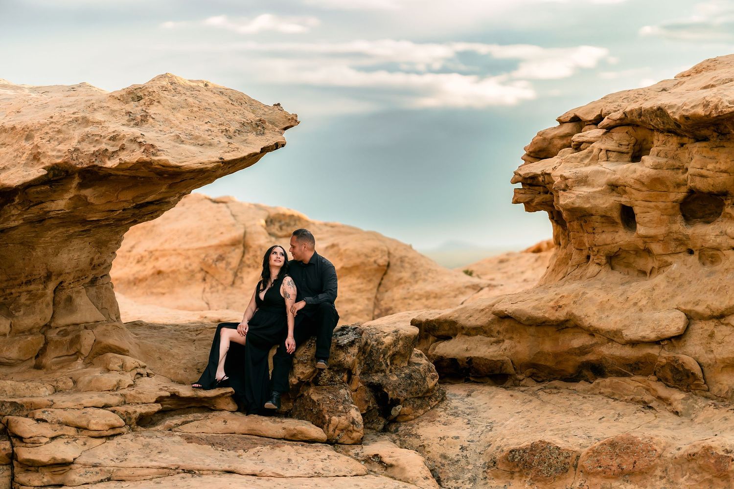 Engaged couple sitting together between sandstone rock formations under a dramatic cloudy sky.