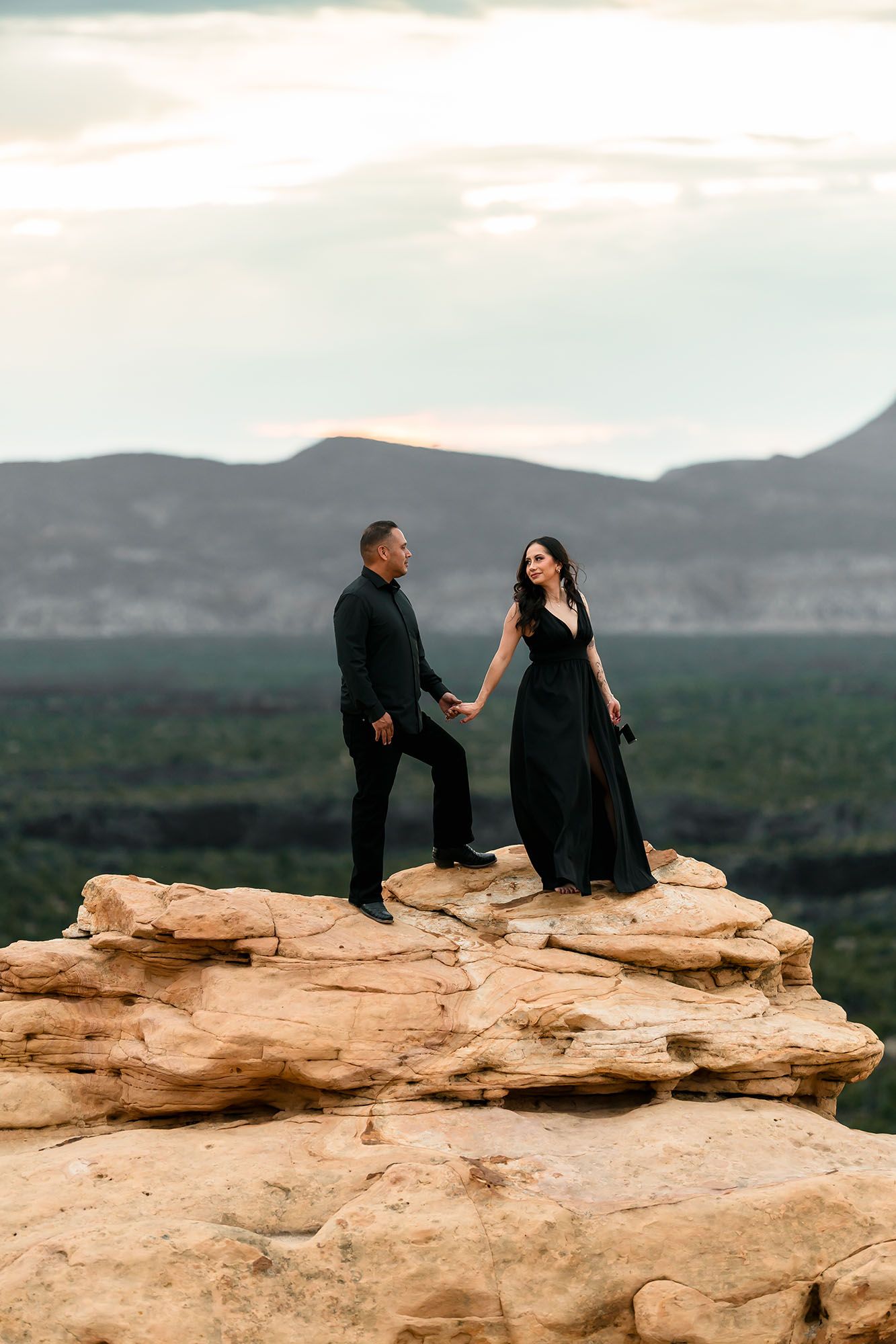 Couple in black holding hands on sandstone summit with mountain and forest backdrop at dusk.