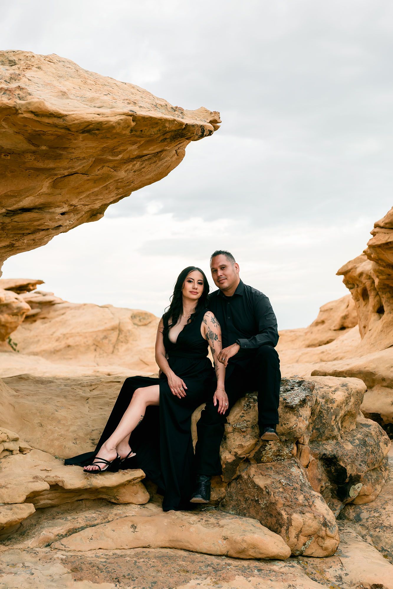 Elegant couple in matching black outfits posing on desert rocks beneath a dramatic sandstone arch.