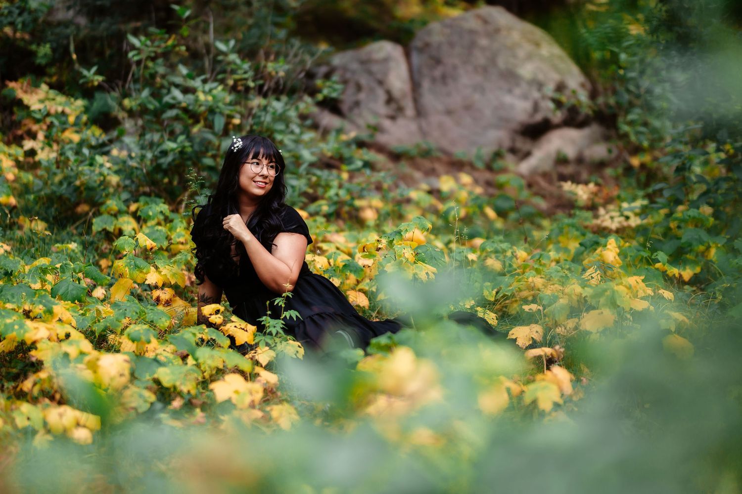 Woman in black dress sits peacefully among yellow and green foliage near large rocks.
