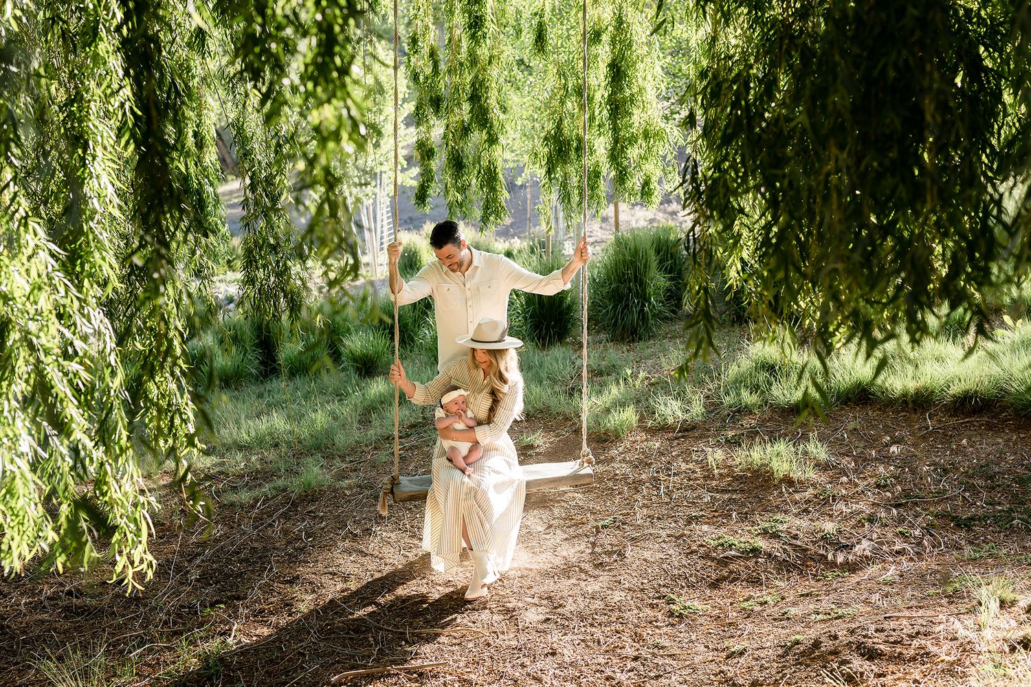 Family enjoying a wooden swing under weeping willow trees in a sunlit garden setting.