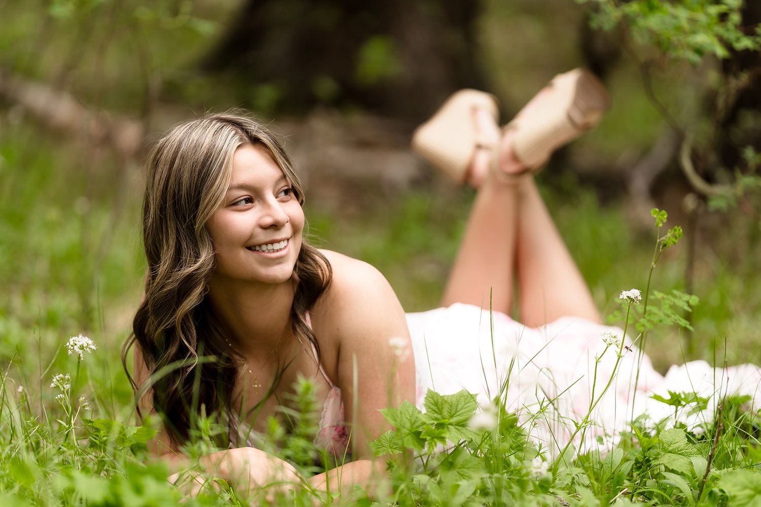Smiling young woman lying in green grass and wildflowers, enjoying a sunny day outdoors in a white dress.