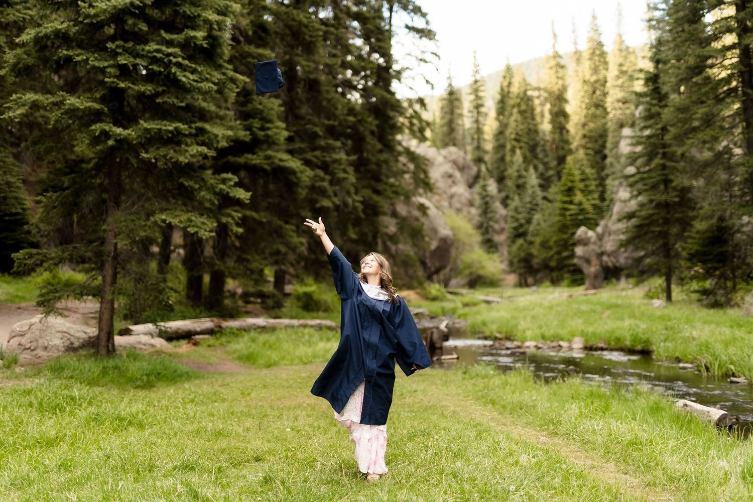 Graduate in navy gown tosses cap in lush green mountain meadow surrounded by tall pine trees.