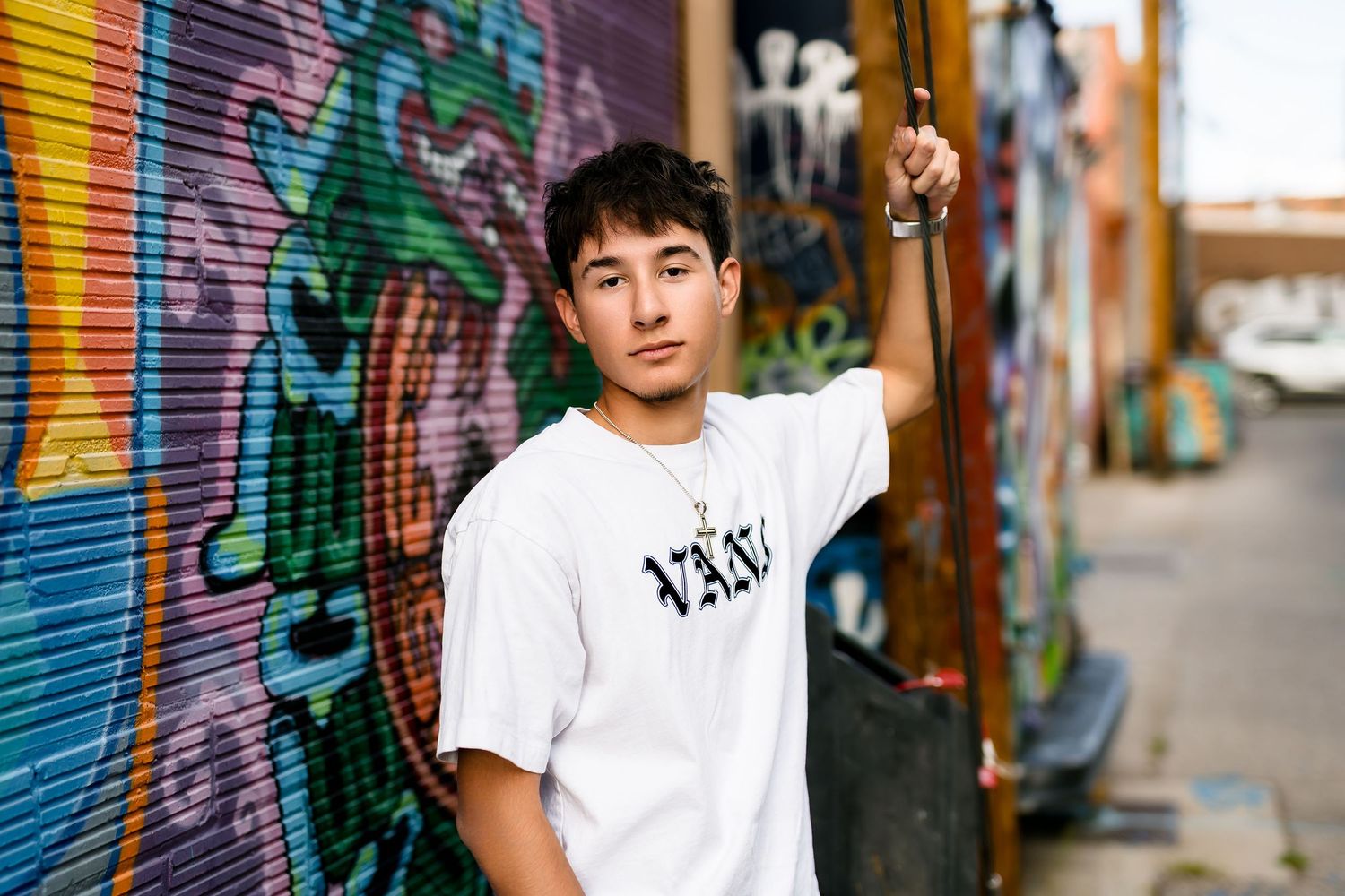 Young man in white Vans tee leaning against vibrant graffiti wall in urban alley.