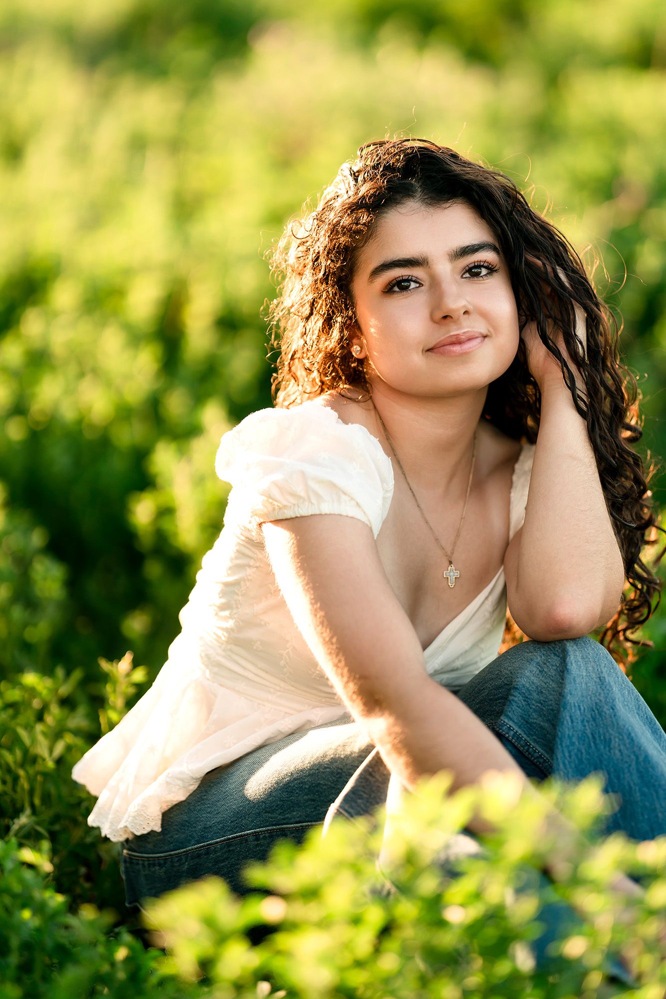 Young woman with curly hair sitting in green field, wearing white top and jeans, smiling softly in golden sunlight.