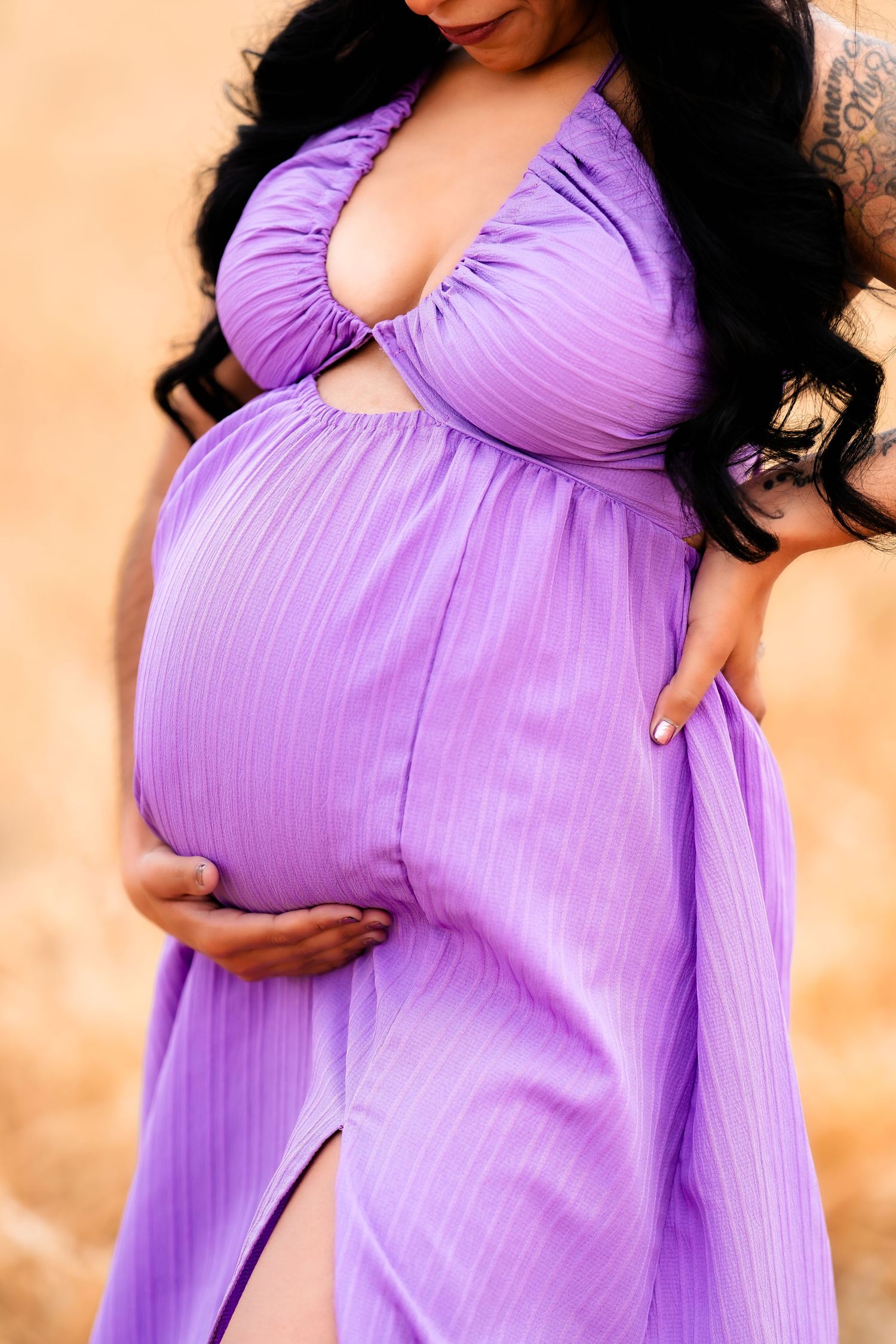 A pregnant figure wearing a flowing purple maternity dress with a plunging neckline cradles their belly against a neutral background.