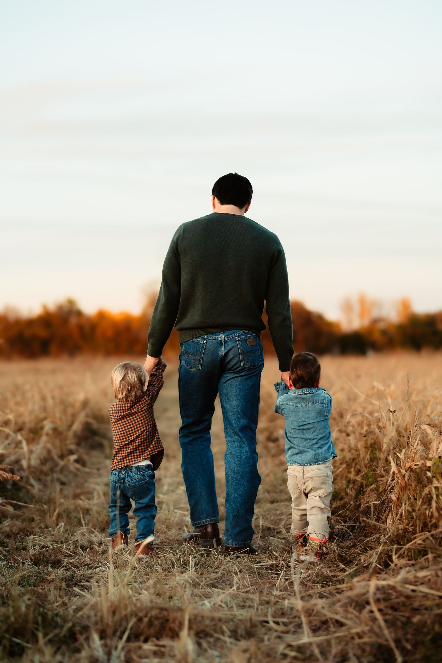 A person in a dark sweater walks through an autumn field at sunset holding hands with two small children.
