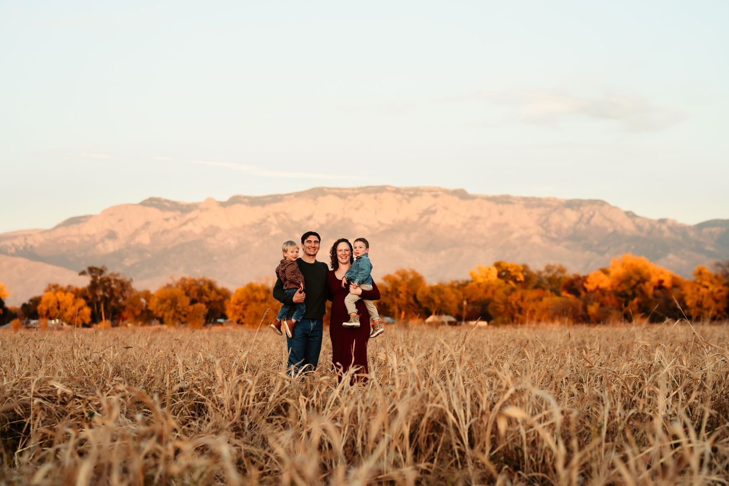 A group stands together in a golden wheat field at sunset with mountains and autumn trees in the background.
