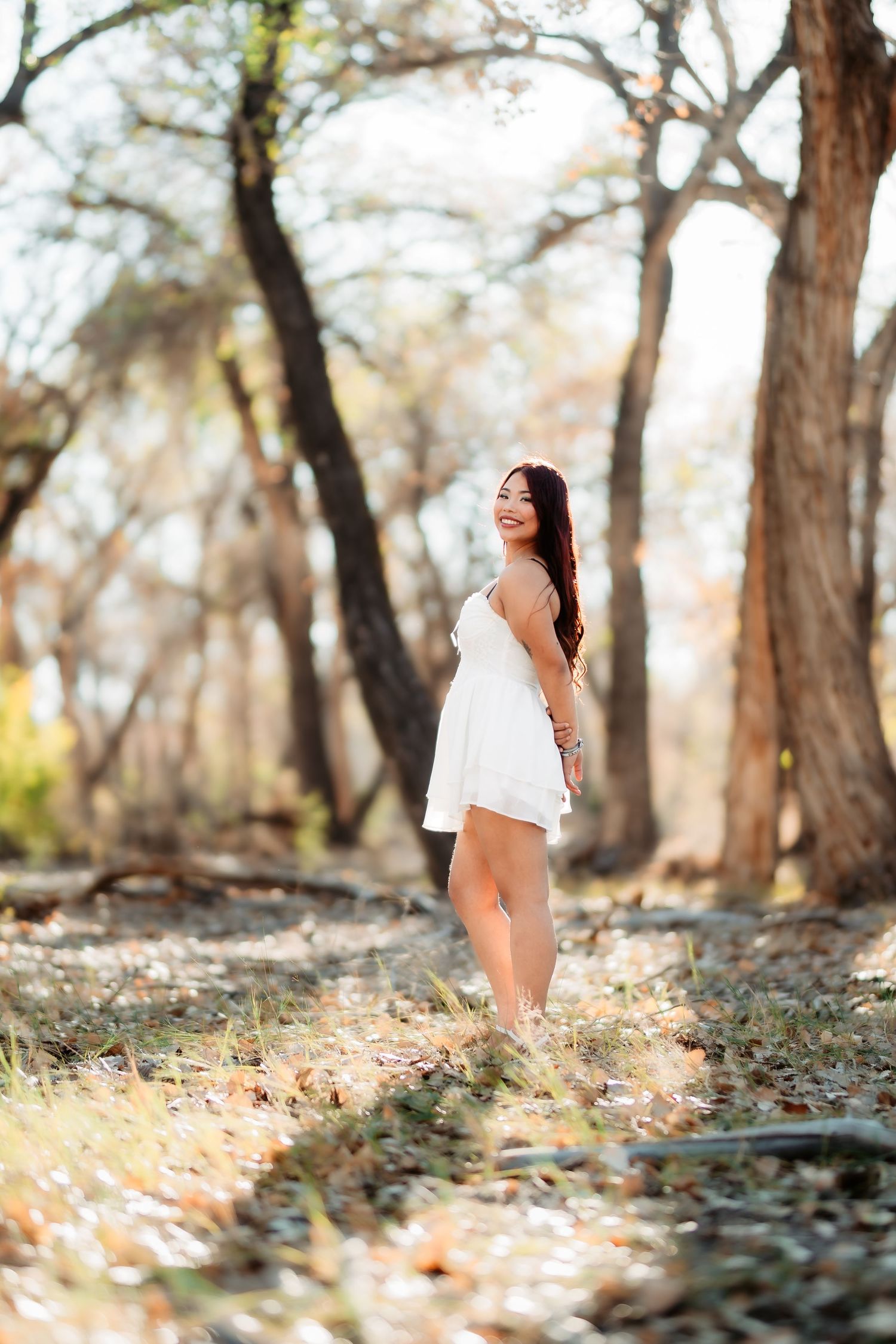 A woman in a white dress poses playfully in a sunlit wooded area with autumn leaves on the ground.
