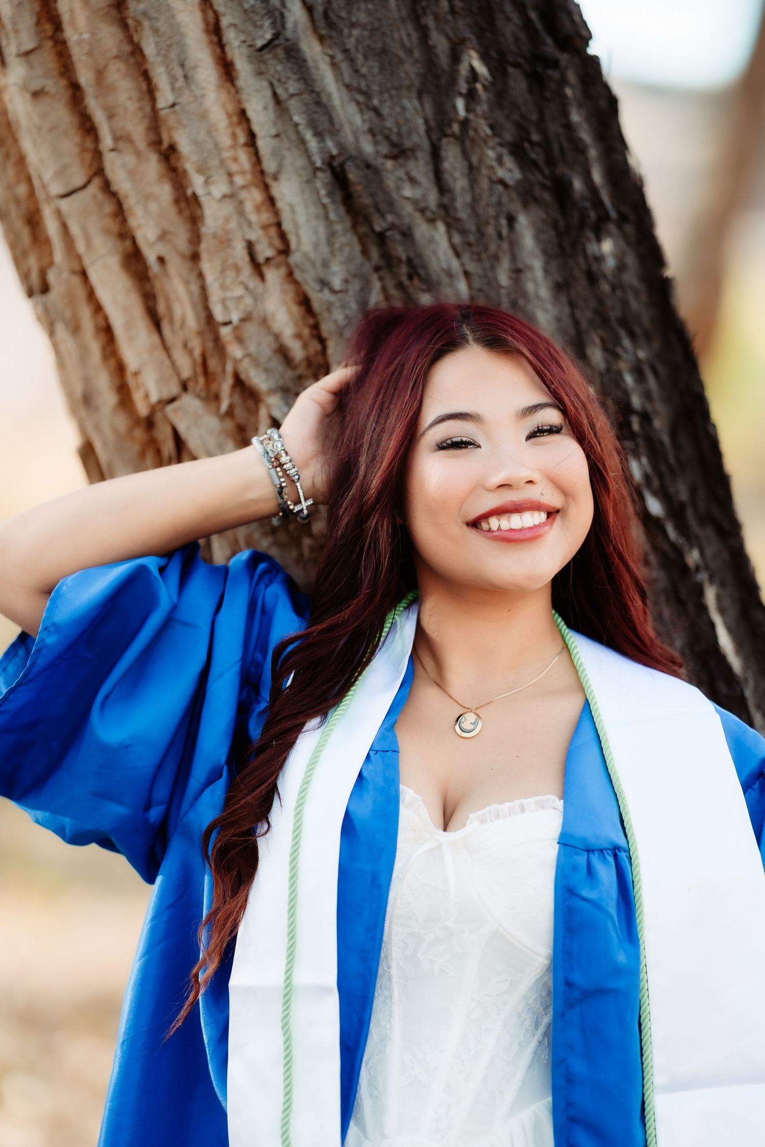 Graduate in blue cap and gown smiles brightly while posing against tree trunk backdrop.