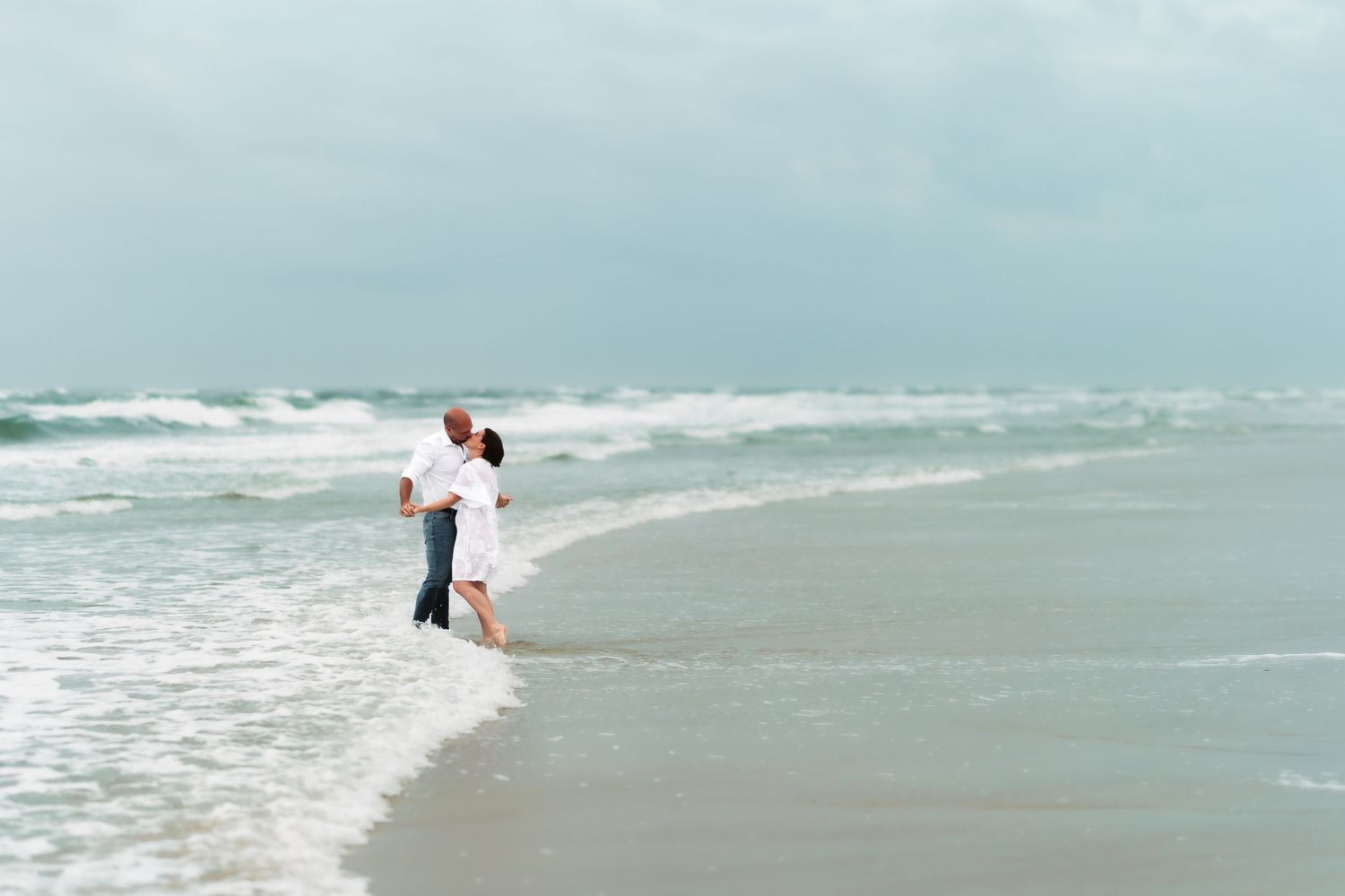 A couple stands together at the edge of the ocean waves on a misty beach.