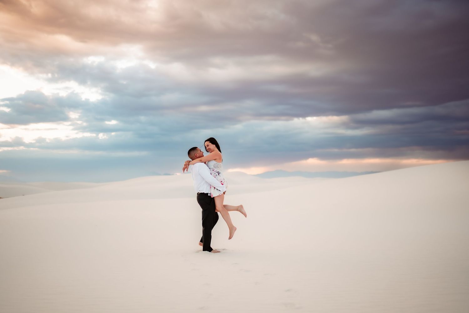 Romantic couple embraces on a beach at sunset with dramatic cloudy sky in the background.