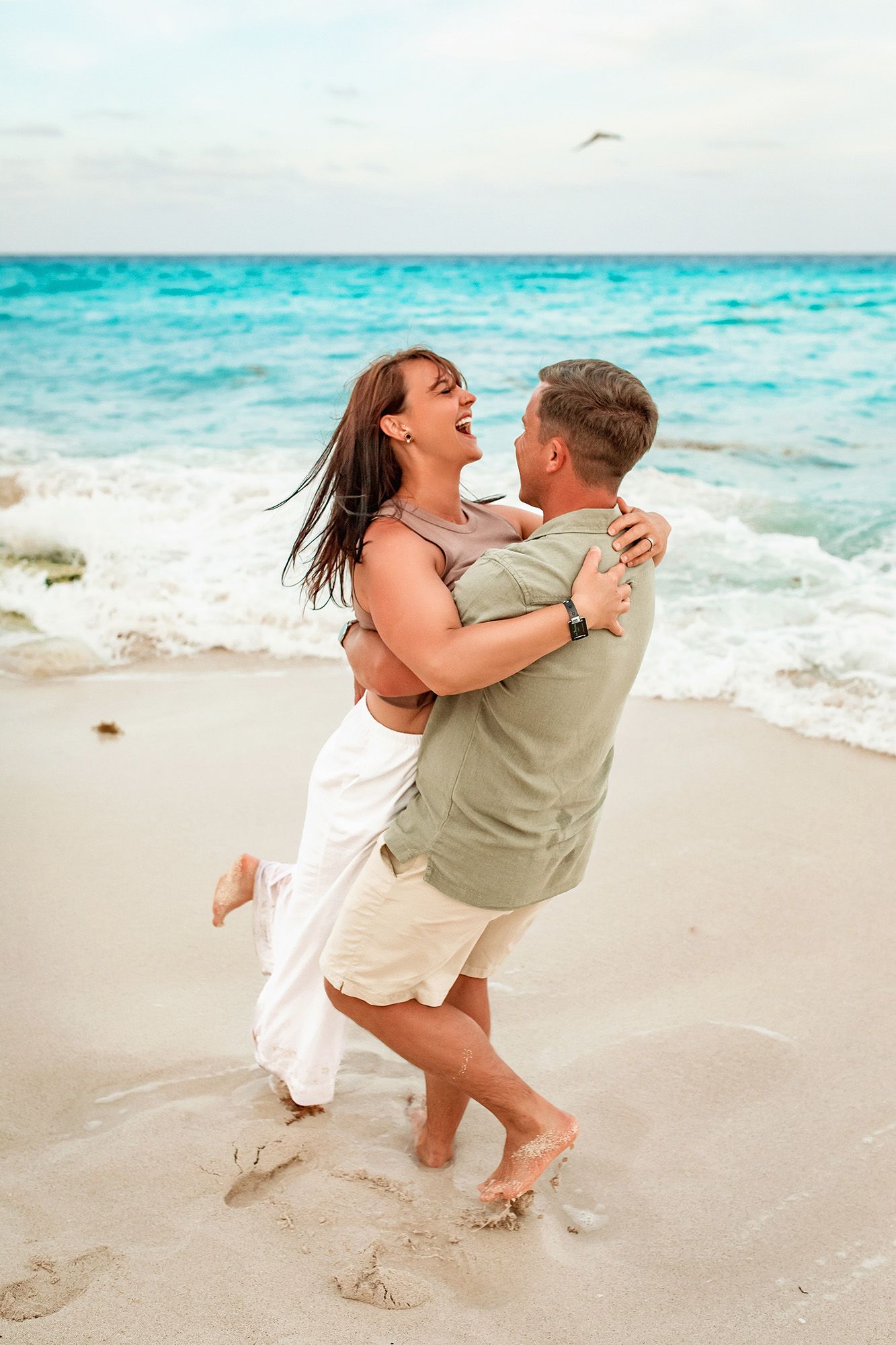 A couple embraces joyfully on a pristine beach with turquoise waters and white sand in the background.