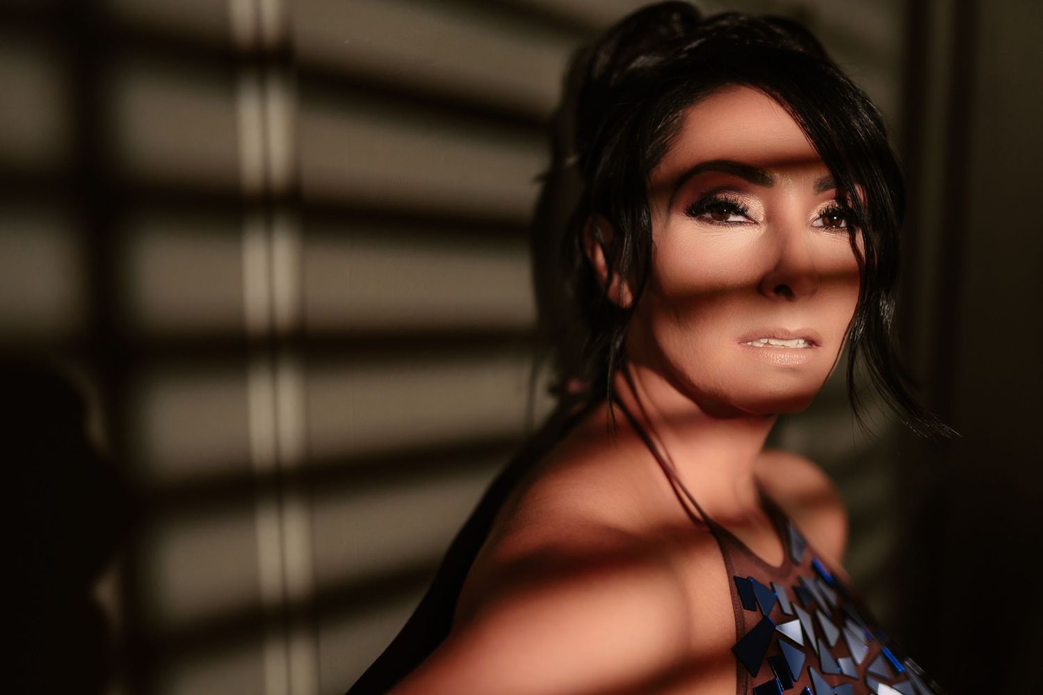 Artistic portrait with dramatic window blind shadows creating stripes across a person's face in moody lighting.
