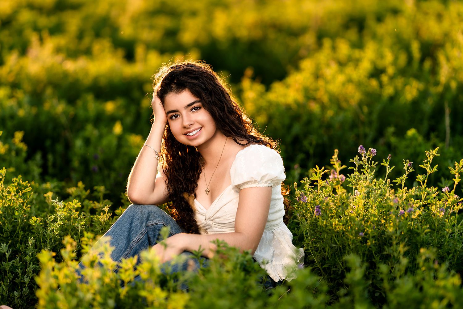 Young woman in white dress sitting among vibrant yellow and purple wildflowers, smiling warmly in golden sunlight.