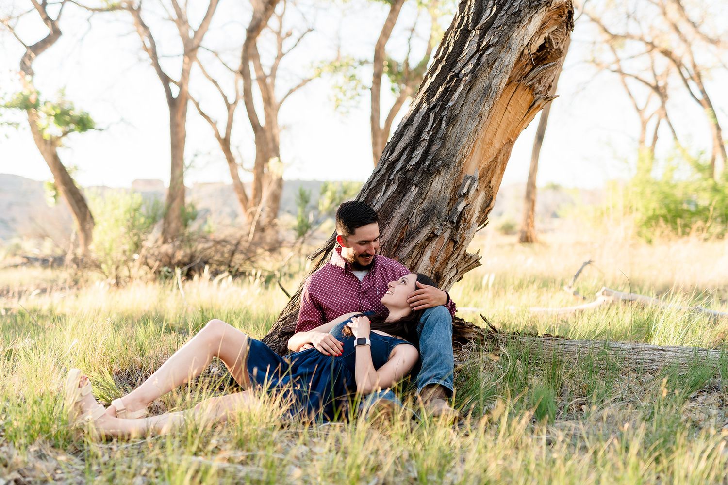 Couple sitting together against a tree trunk in a sunny field, sharing a romantic moment outdoors.