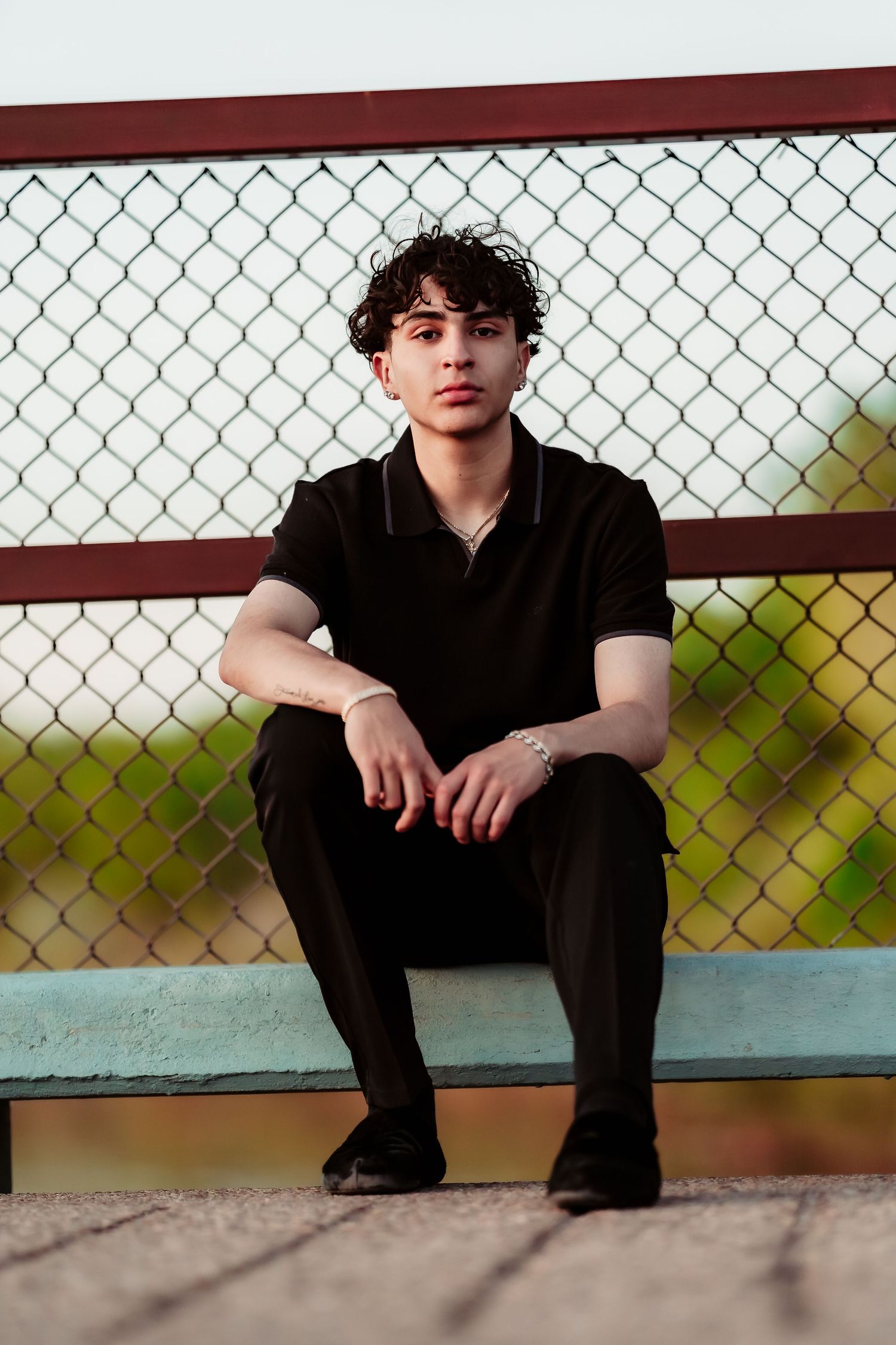 Young person in black outfit sitting on bench against chain link fence during golden hour.