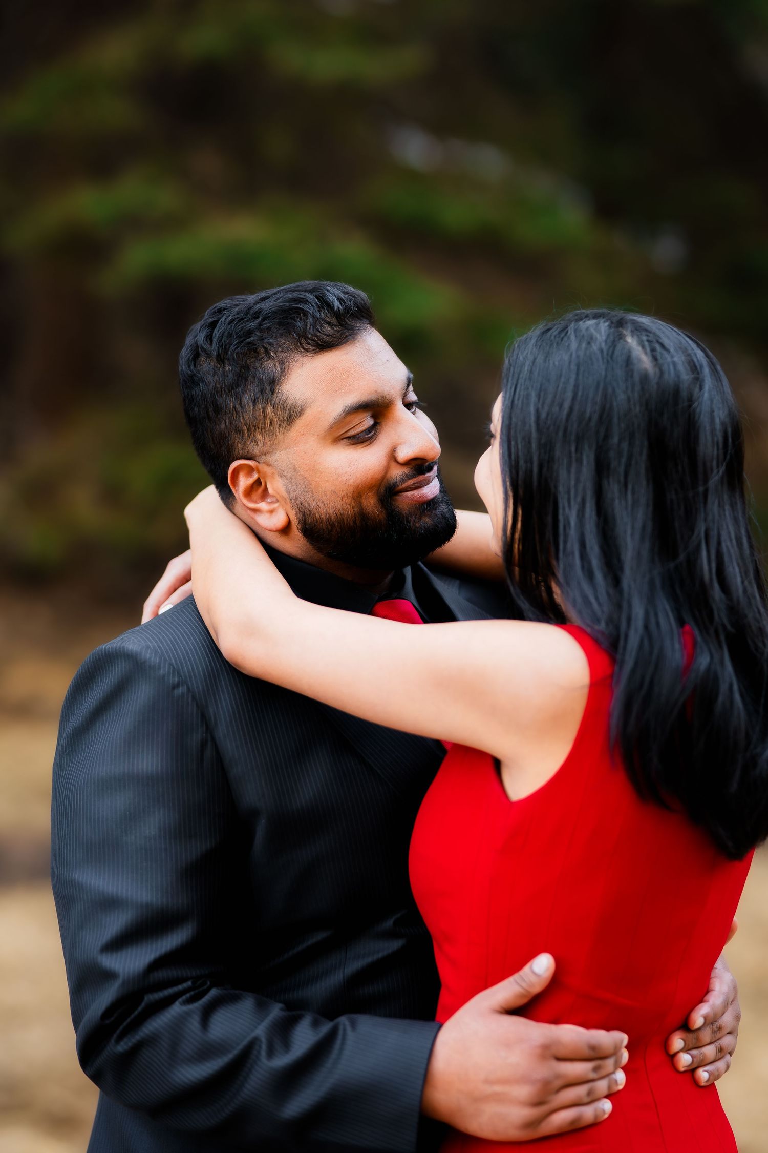 A romantic couple embraces outdoors, with one wearing a red dress and the other in a black suit.
