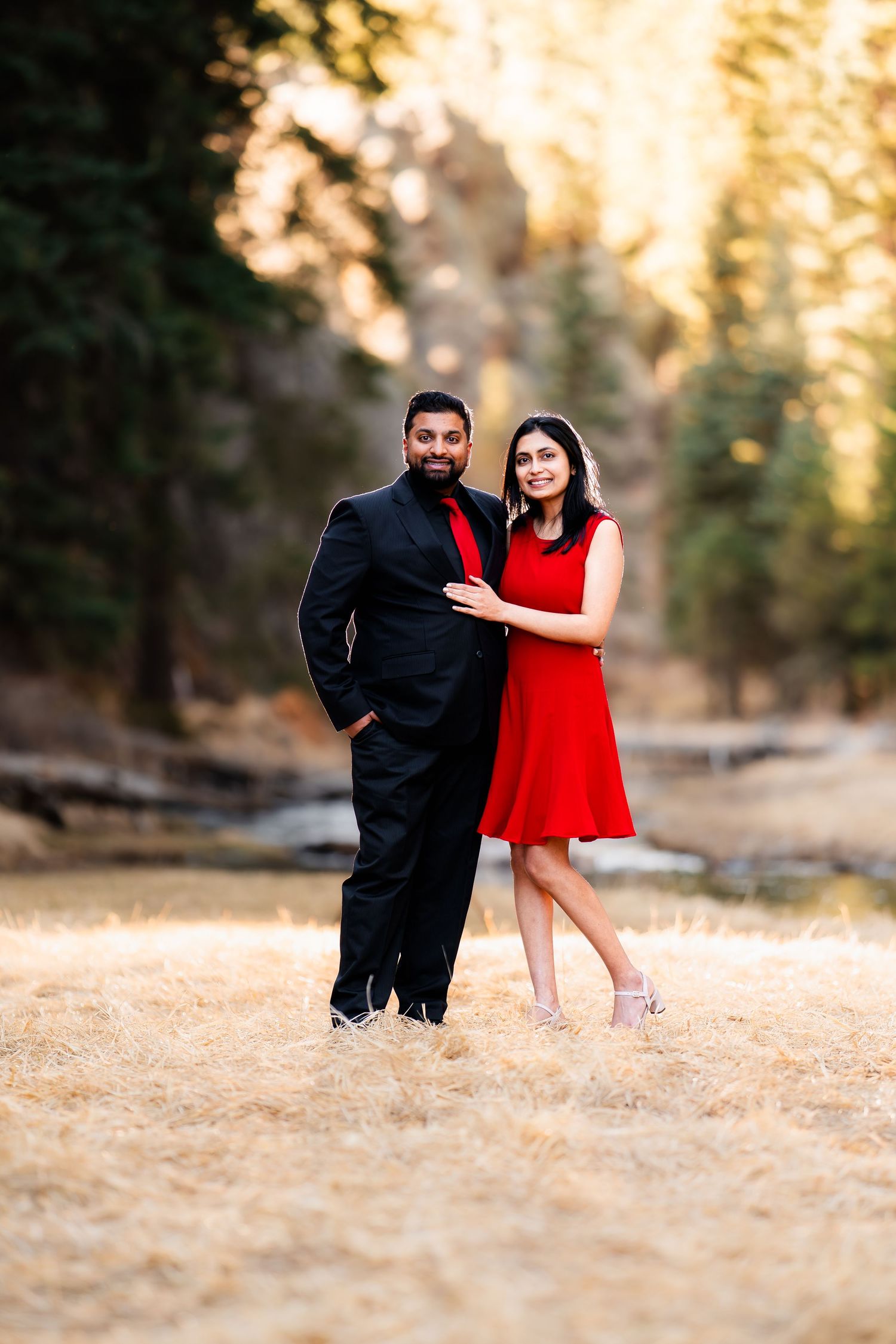 A couple in formal attire poses outdoors, with the person in red dress and person in black suit standing together.
