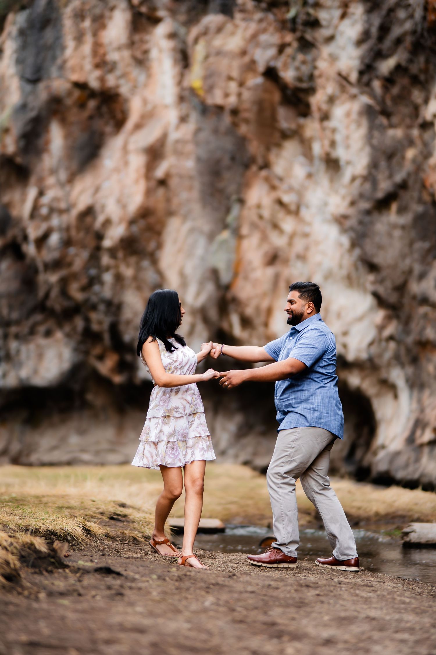 A couple dancing and holding hands in front of a rocky cliff face in casual summer outfits.
