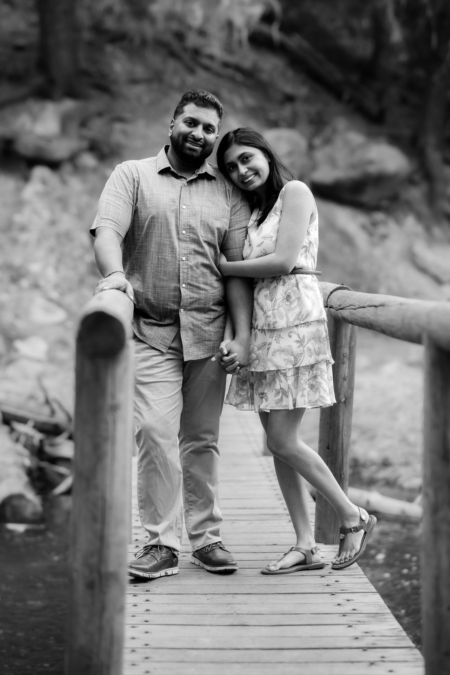 A couple in casual wear poses together on a wooden boardwalk with rocky cliffs behind them.