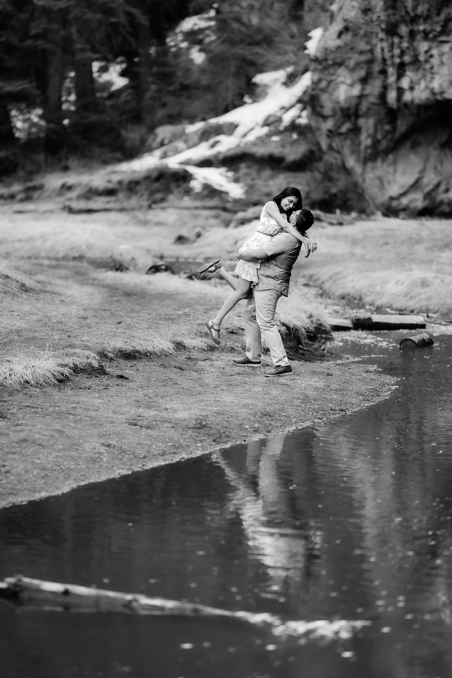 A joyful couple embraces by a reflective pond with rocky cliffs in the background.