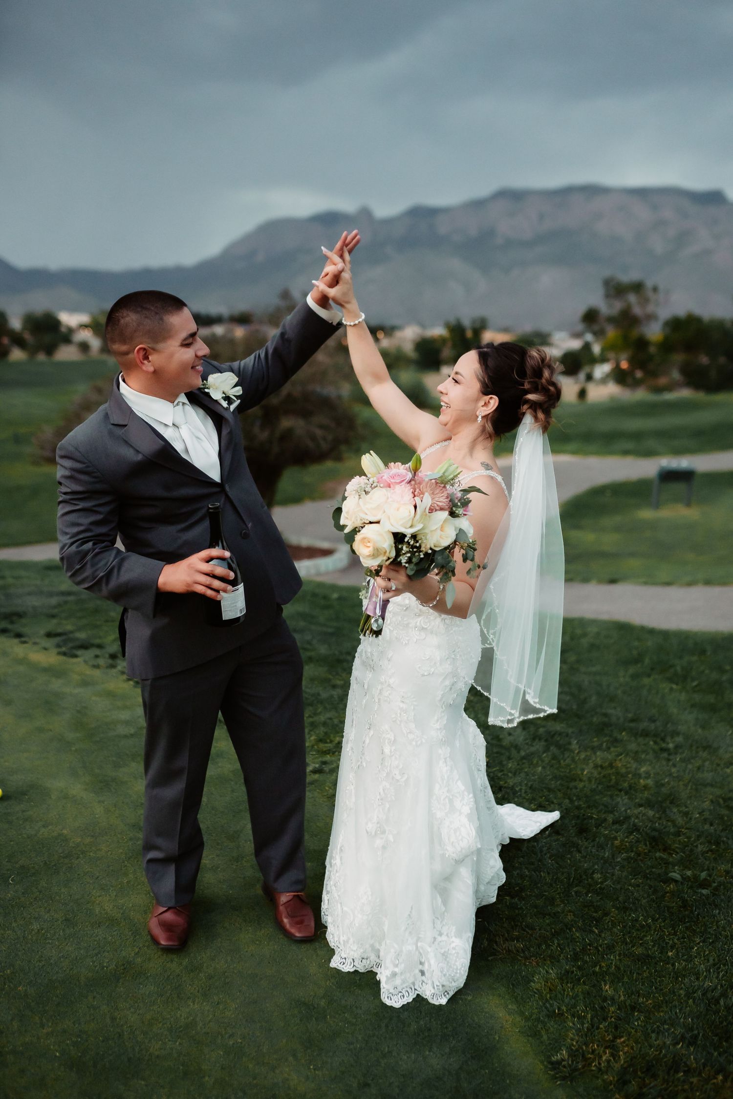 Playful wedding couple dances and dips on grassy hillside with mountains in background.