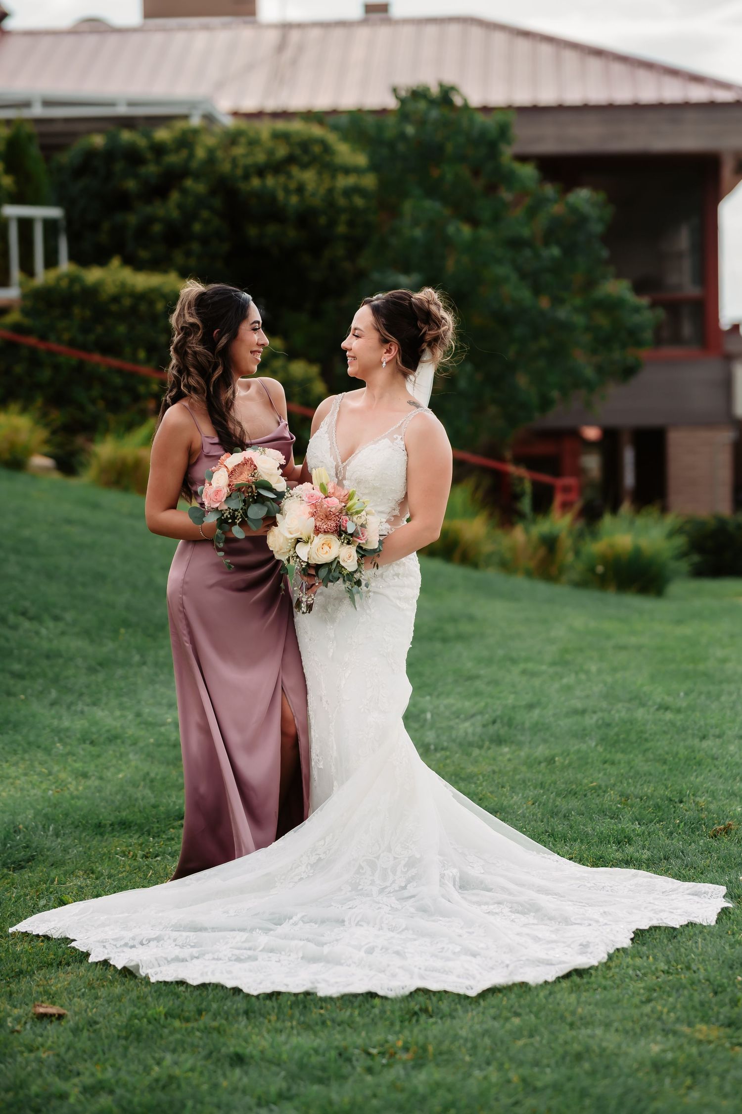 A bride in a flowing white lace gown sits gracefully on green grass during a wedding photoshoot.