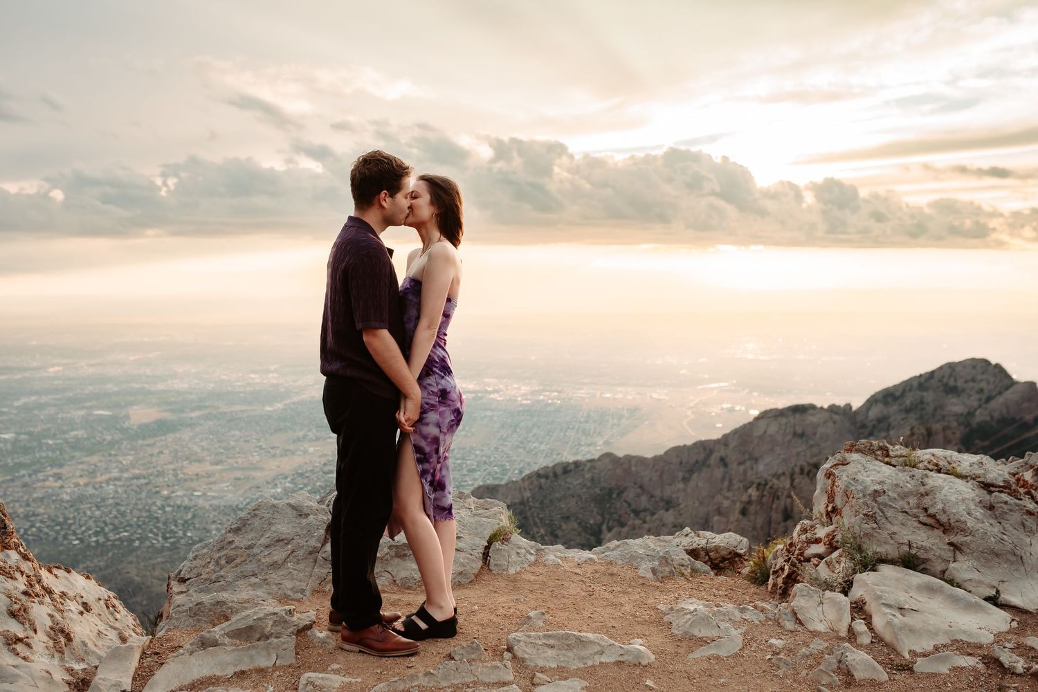 Romantic couple shares intimate moments on a mountain ridge during sunset with dramatic sky and mountains in the background.