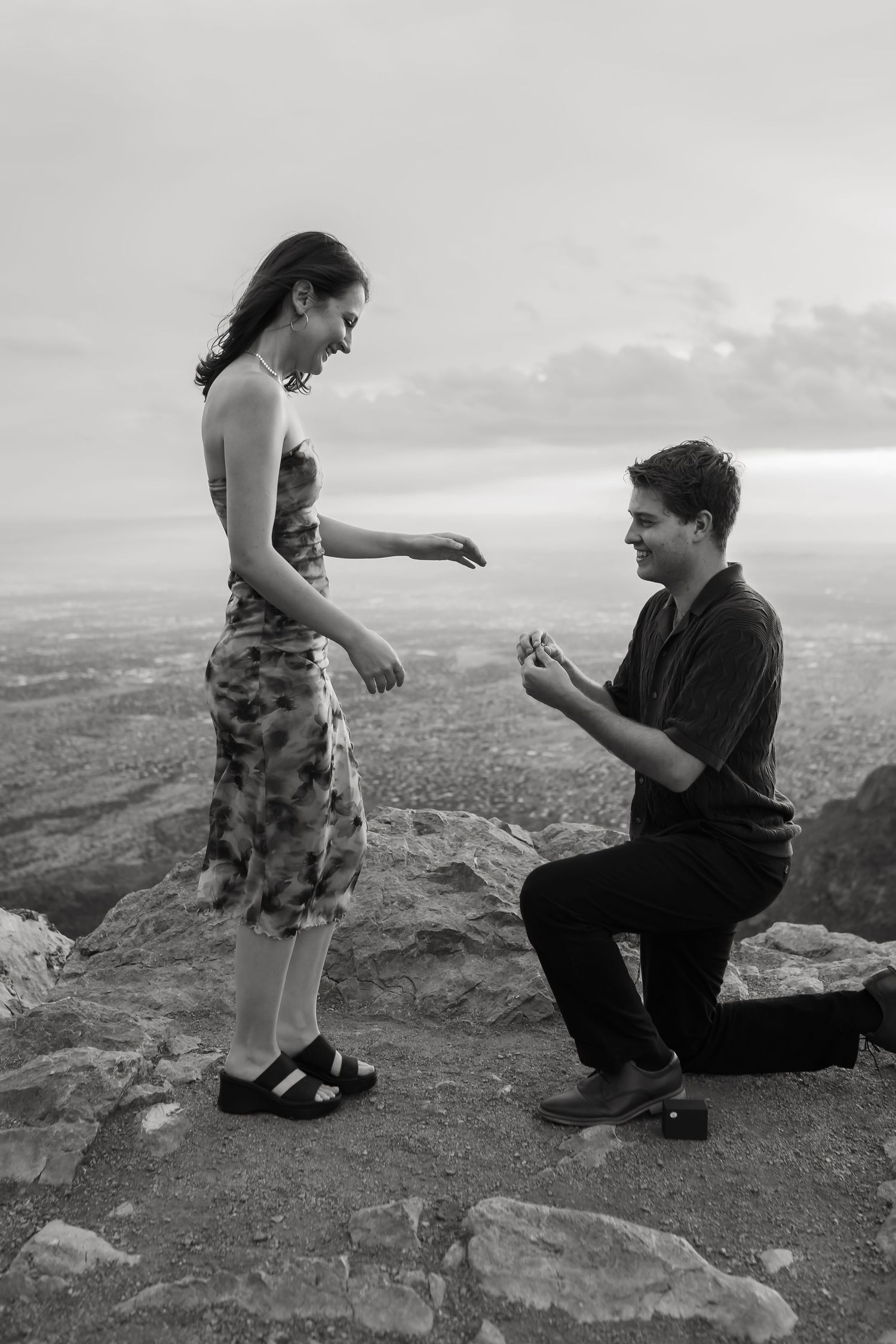 A proposal moment captured in black and white on a dramatic mountain cliff overlook.