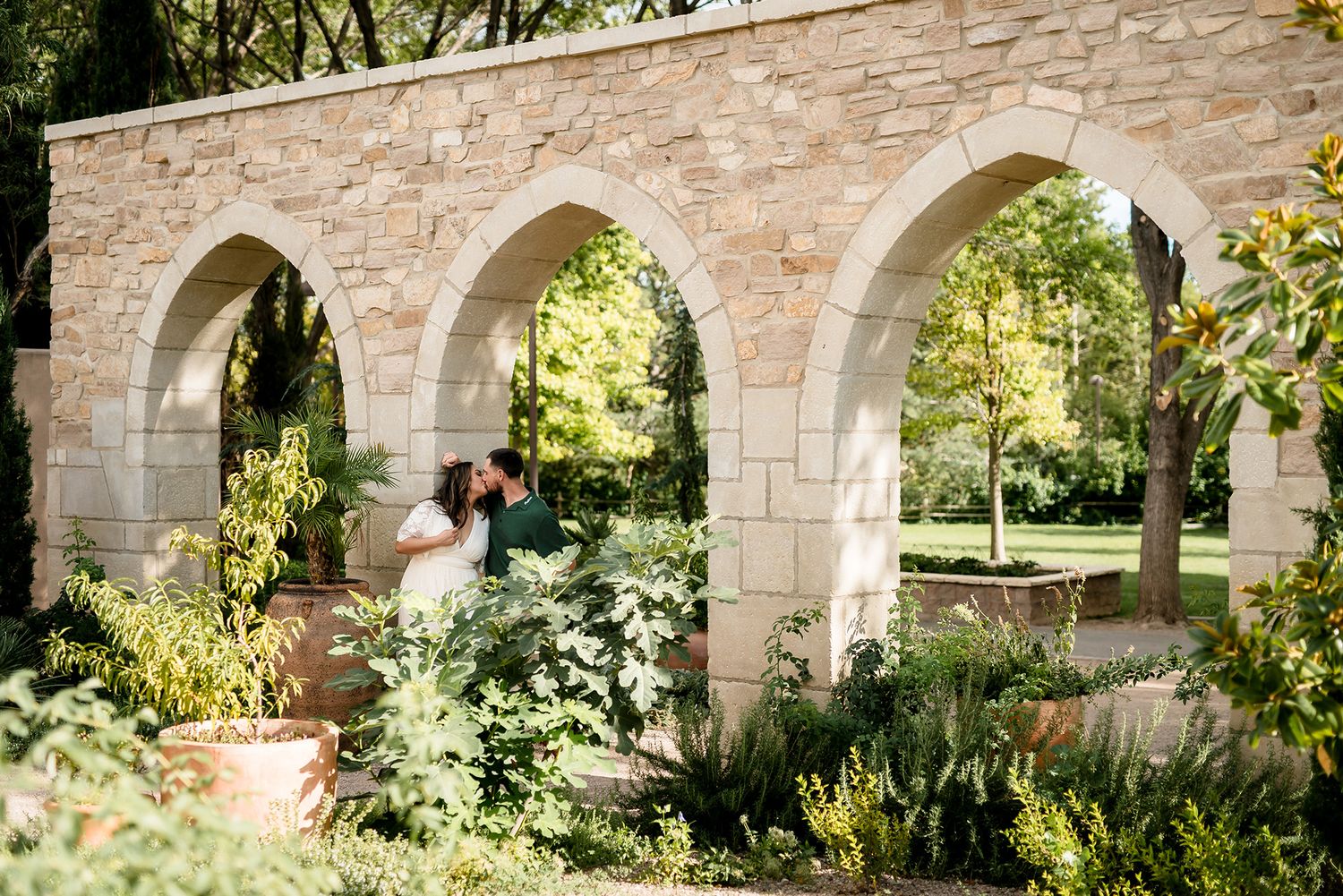 Couple sharing a romantic kiss under stone arches surrounded by lush garden greenery in an elegant outdoor setting.