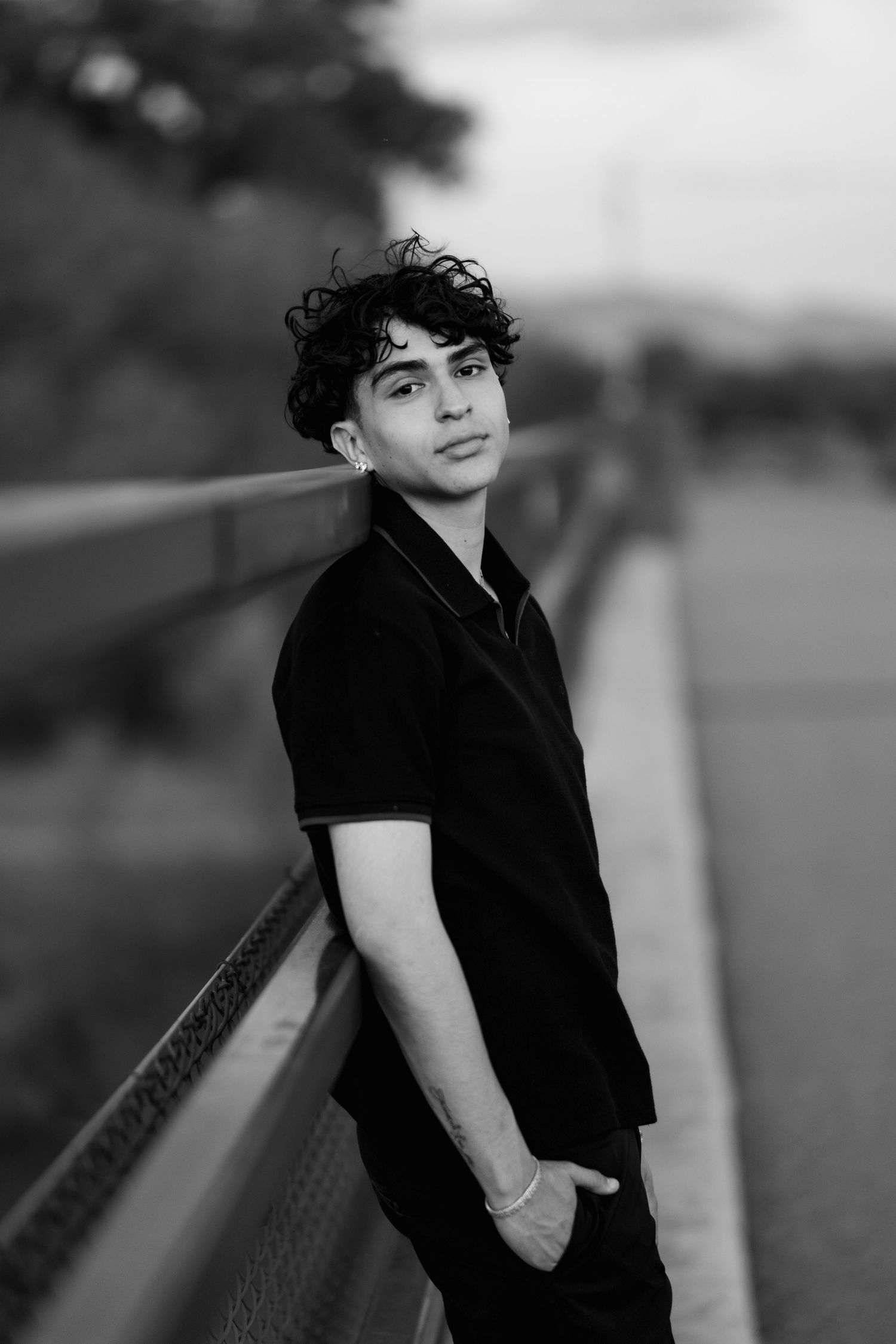 Black and white portrait of someone with curly hair in a black polo shirt leaning against a railing outdoors.