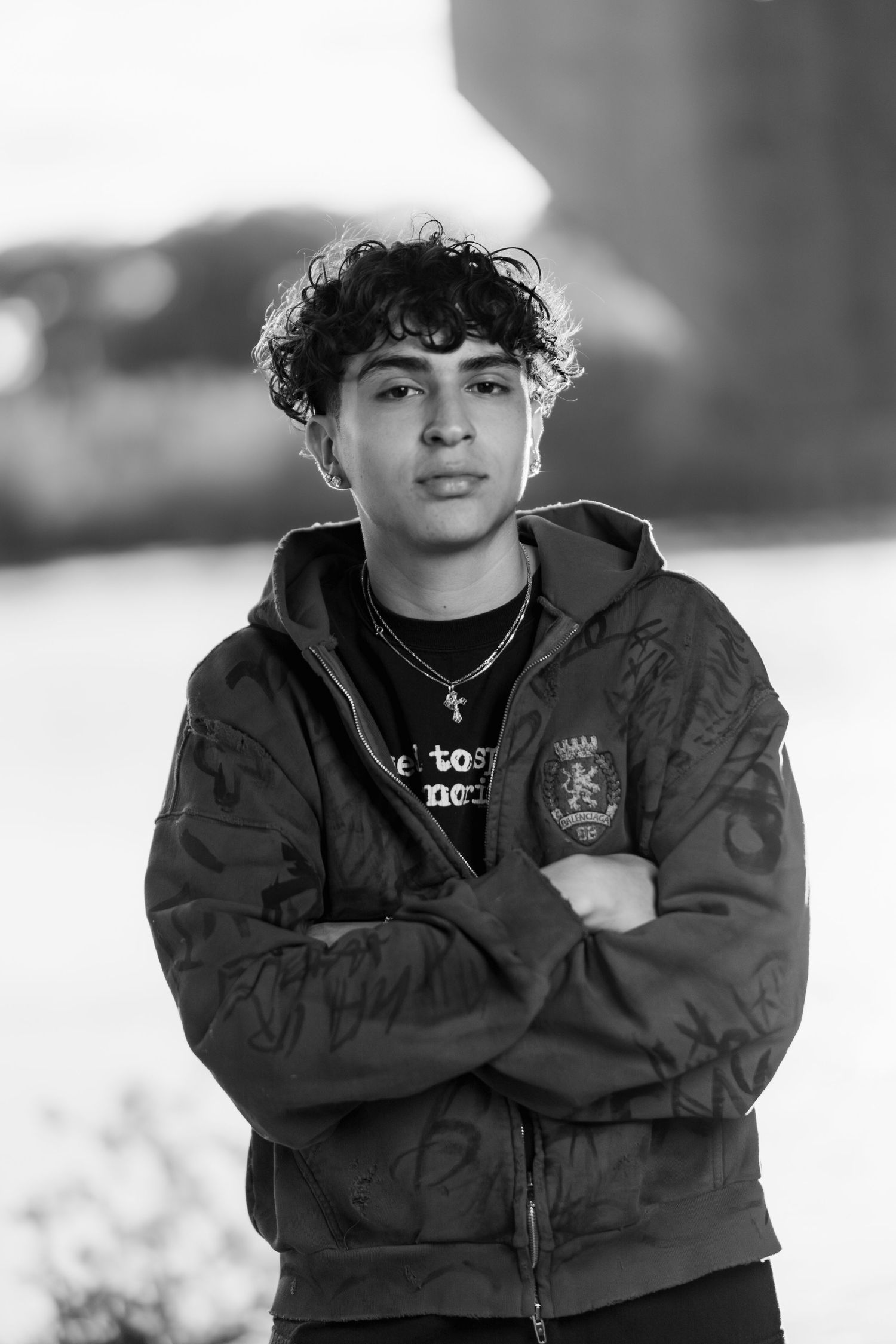 Black and white portrait of a person in a dark bomber jacket standing with arms crossed outdoors on a bright day.