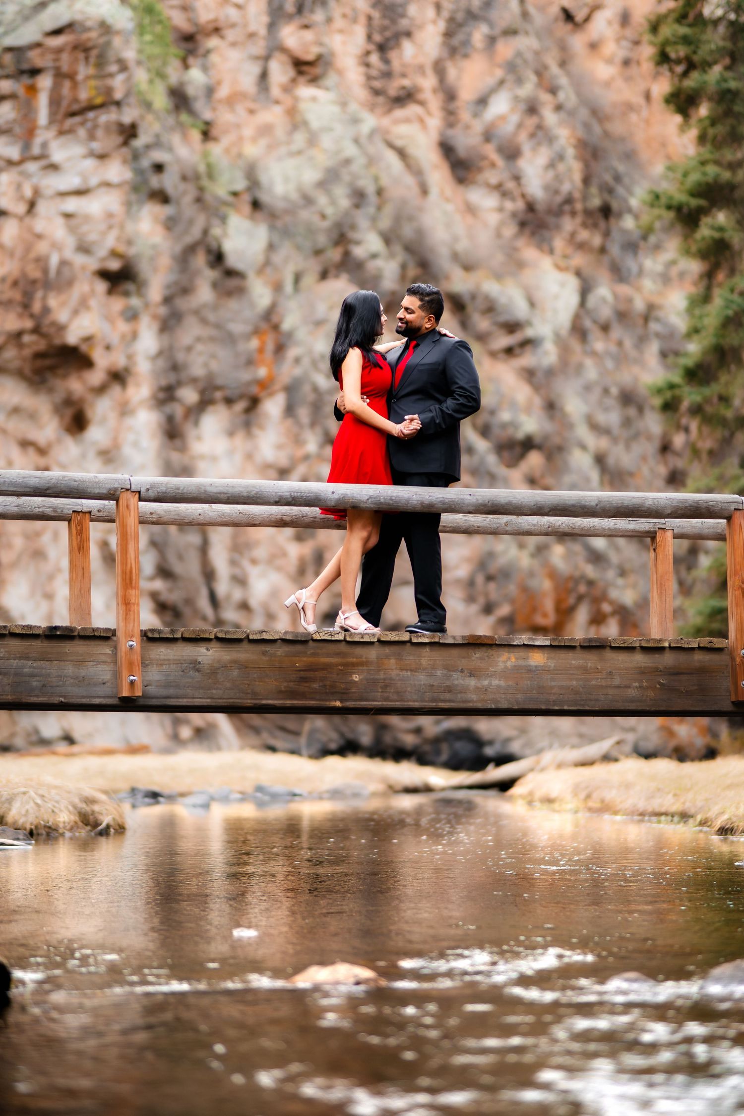 A couple embrace on a wooden footbridge over calm water with dramatic rocky cliffs in the background.