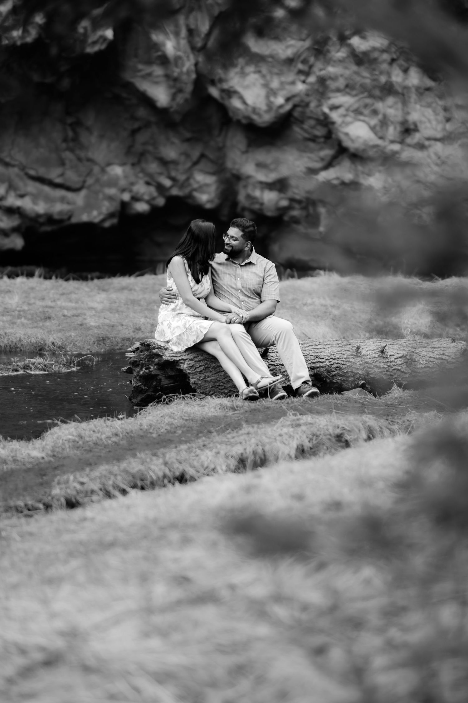 A black and white portrait of a couple sitting by water near rocky cliffs.