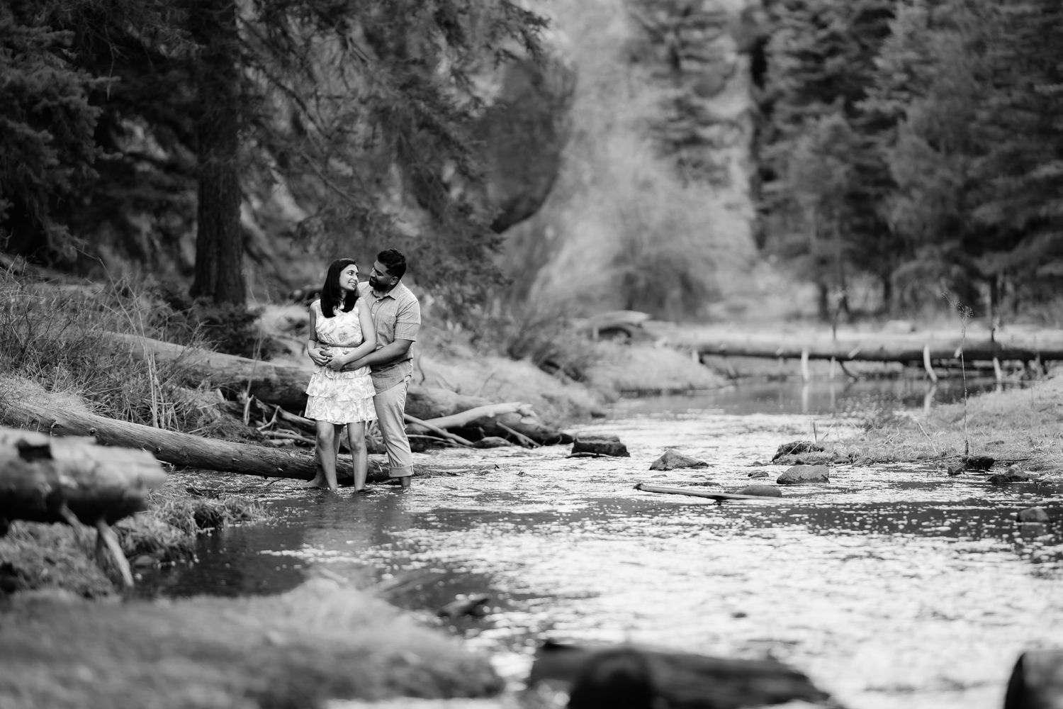 A romantic couple embraces by a stream in a misty forest setting captured in black and white.