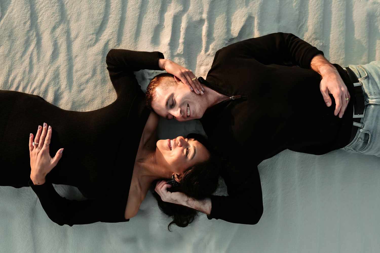 A couple in black clothing lying down together in an artistic pose on a light surface.