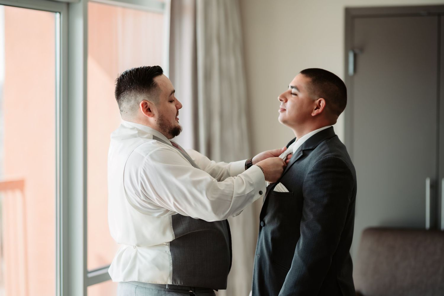 Two men in formal attire having an emotional conversation in a brightly lit room near a window.