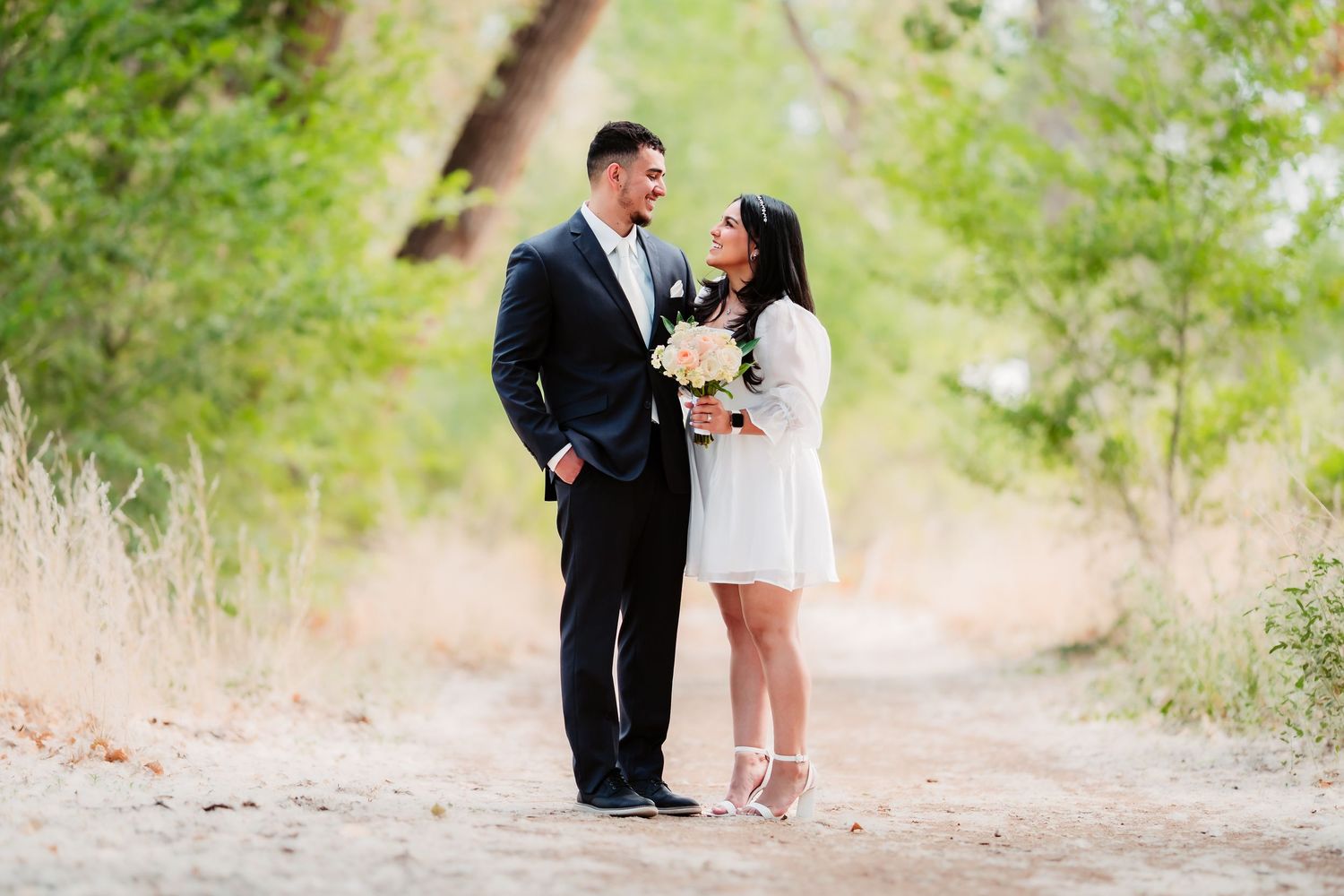 A couple in formal attire shares an intimate moment on a sunlit path surrounded by lush greenery.