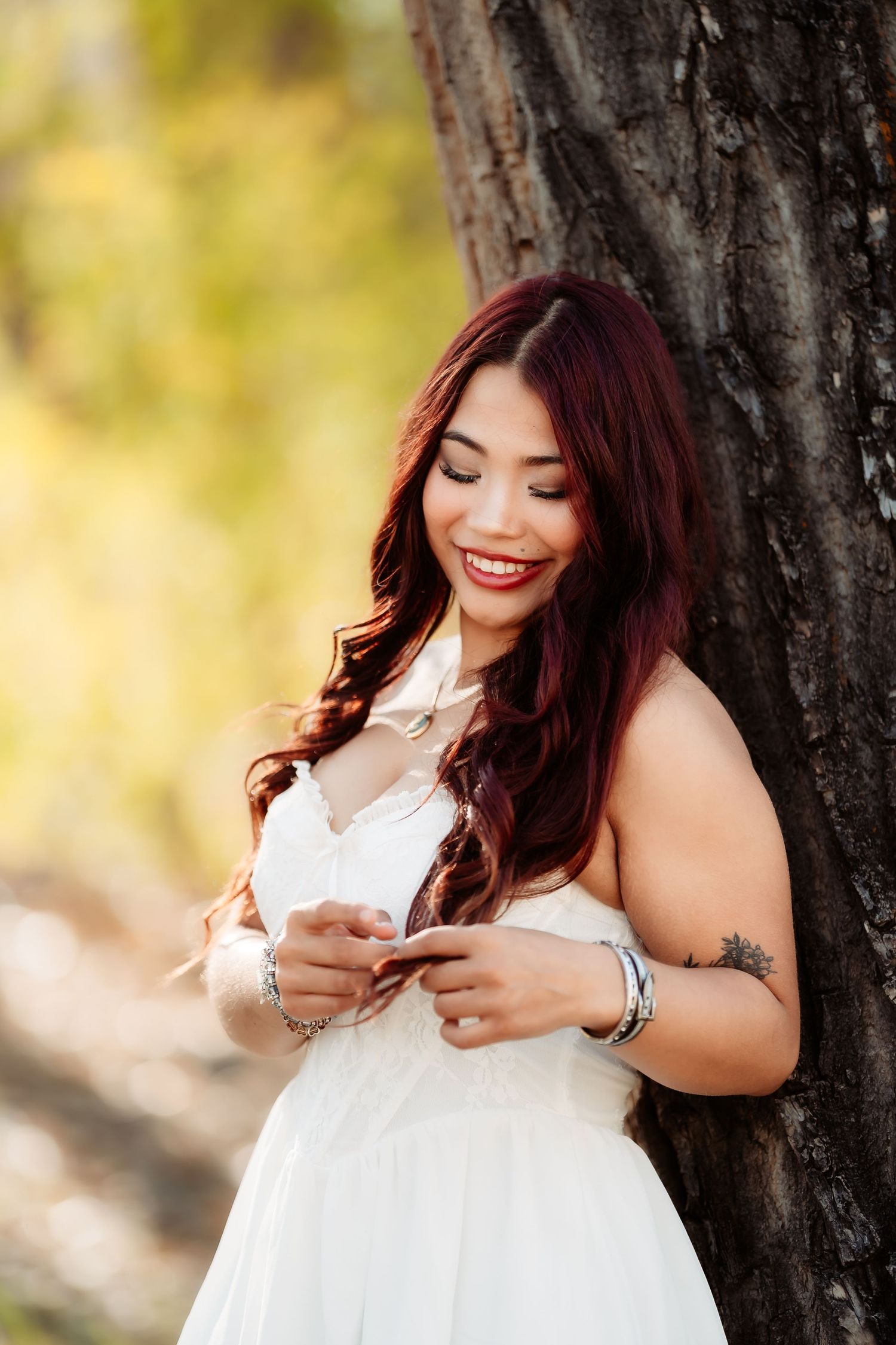 Woman in white dress smiles while posing near tree trunk in outdoor portrait session.