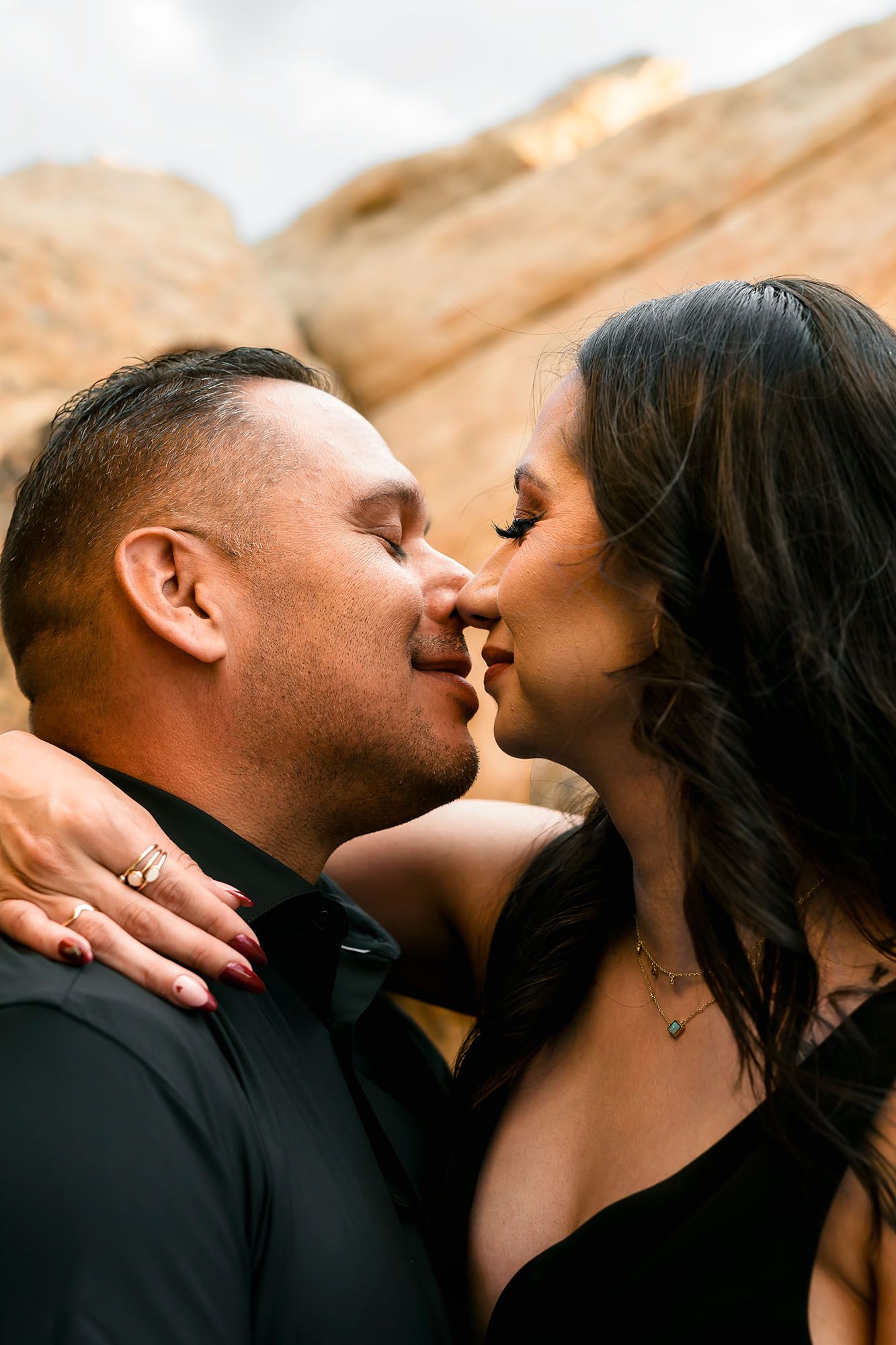 A couple shares a romantic almost-kiss moment against rustic rock formations, both dressed in elegant black outfits.