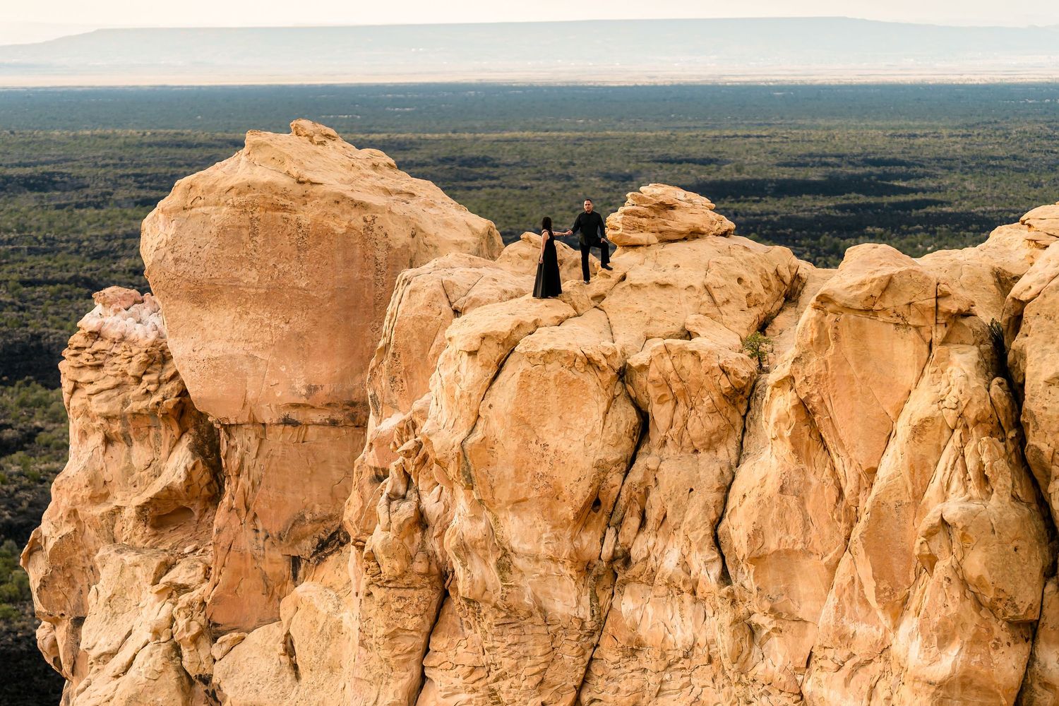 Two people stand atop dramatic golden sandstone rock formations overlooking a vast desert landscape.