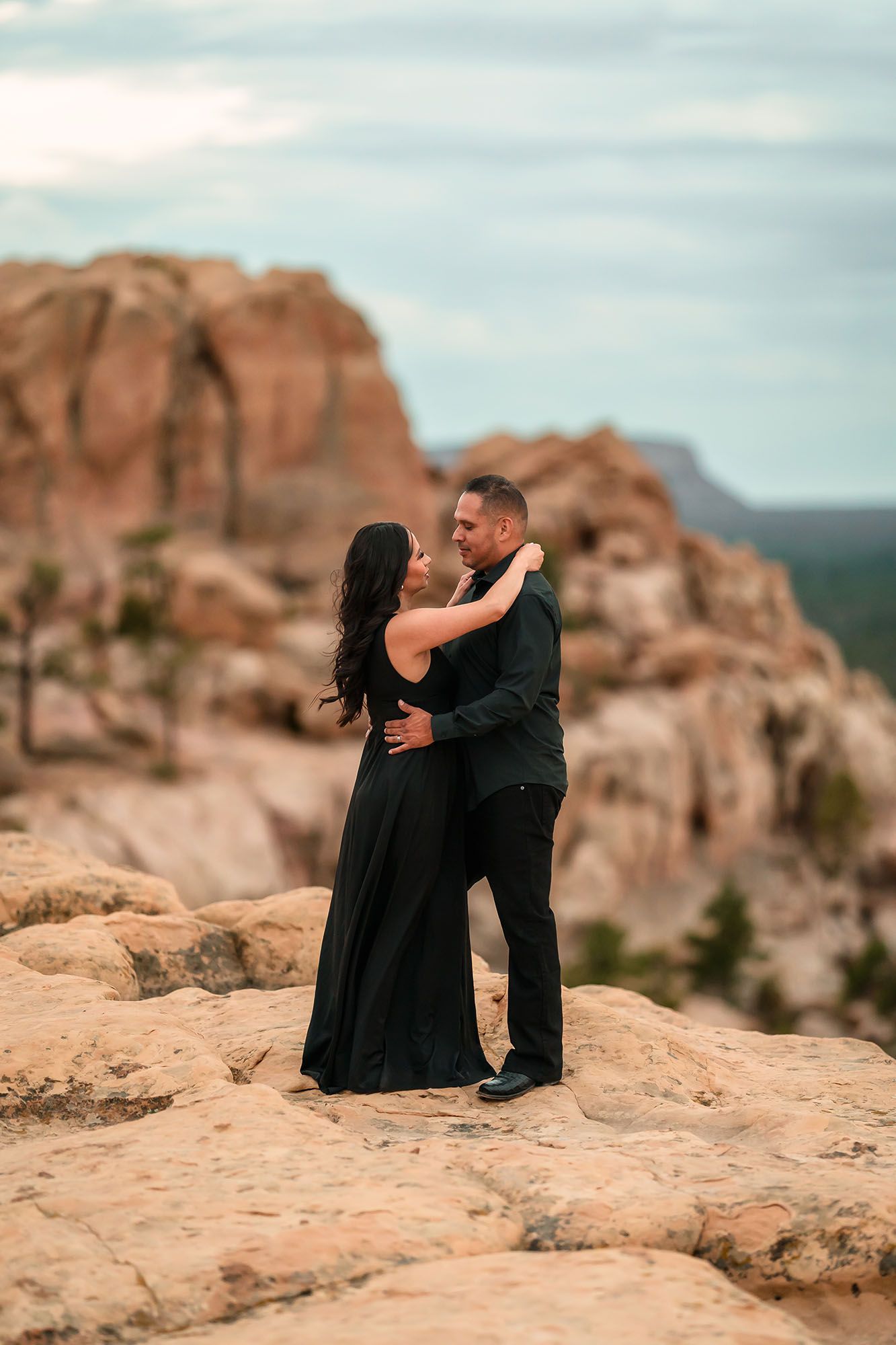 Couple in black formal wear embracing on rocky terrain with red rock canyon backdrop.