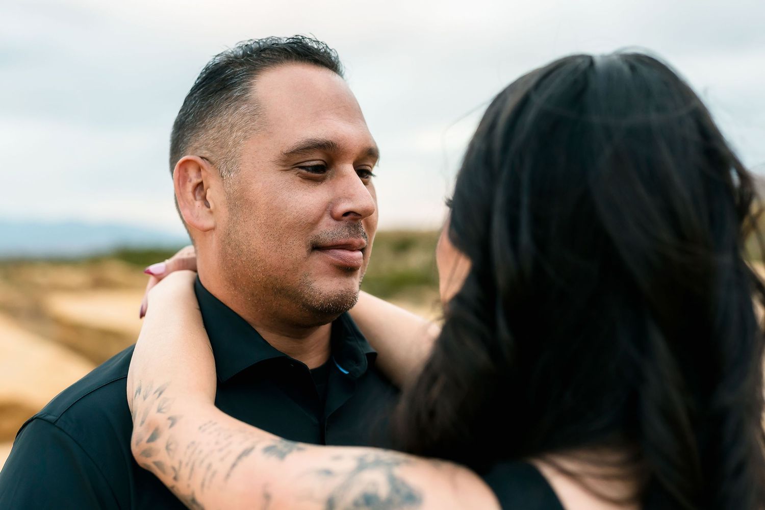 A man with tattoos smiles softly as a woman with dark hair embraces him outdoors in a desert landscape.