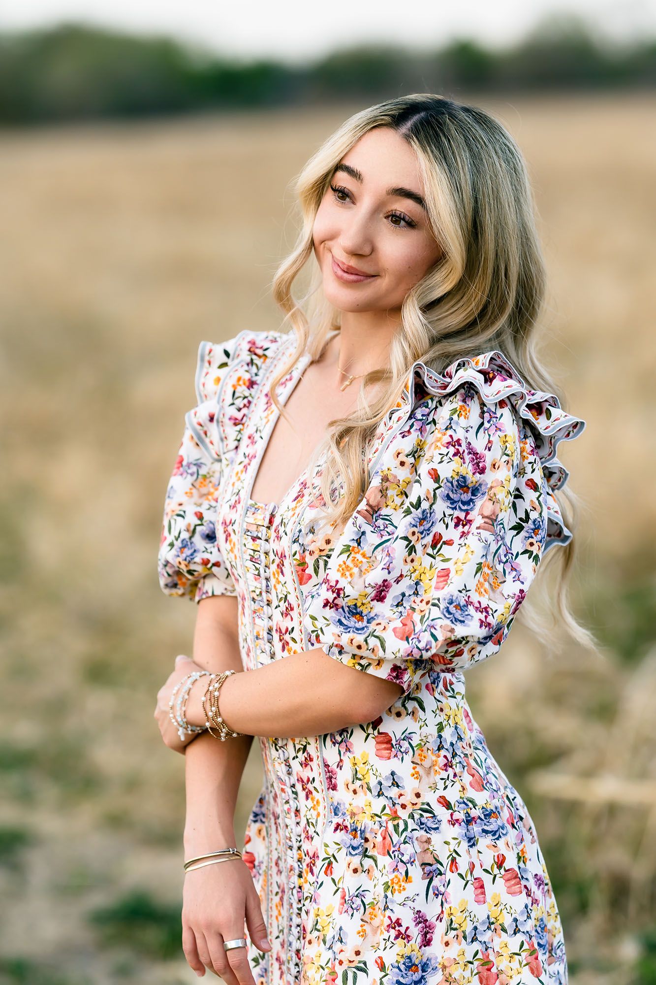 Young blonde woman in floral dress posing in golden field, smiling softly with bracelets and wavy hair.